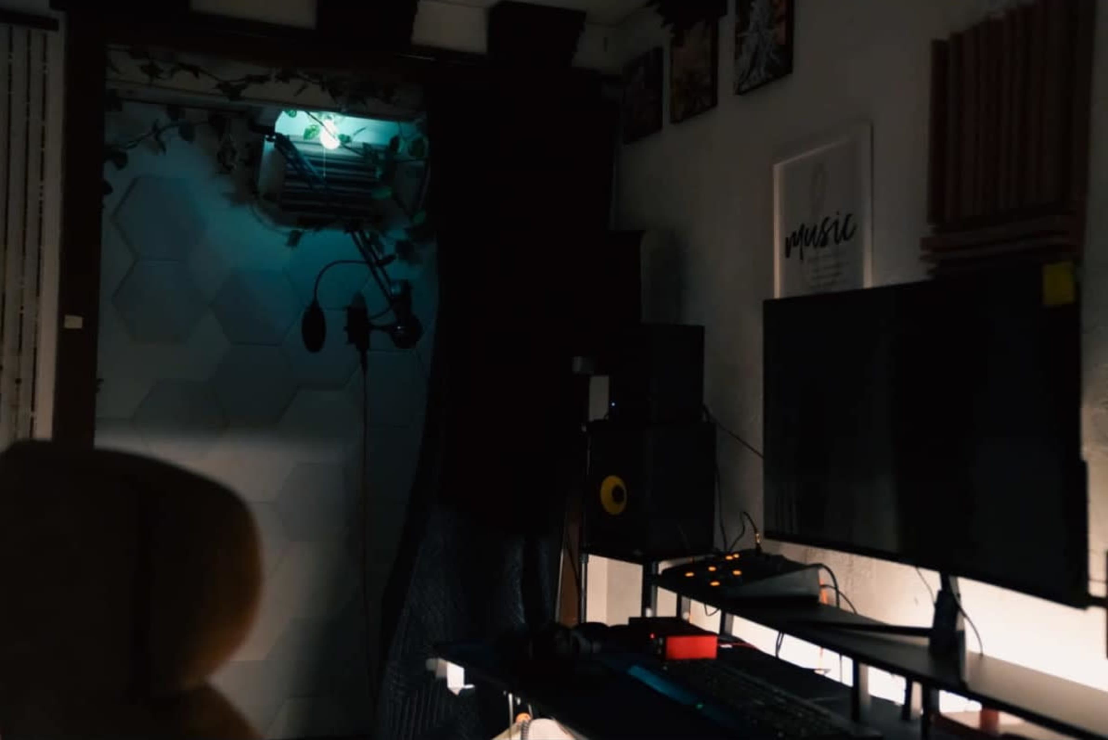 A dimly lit room features a computer desk with monitors and audio equipment, alongside a microphone in front of a partially closed curtain.