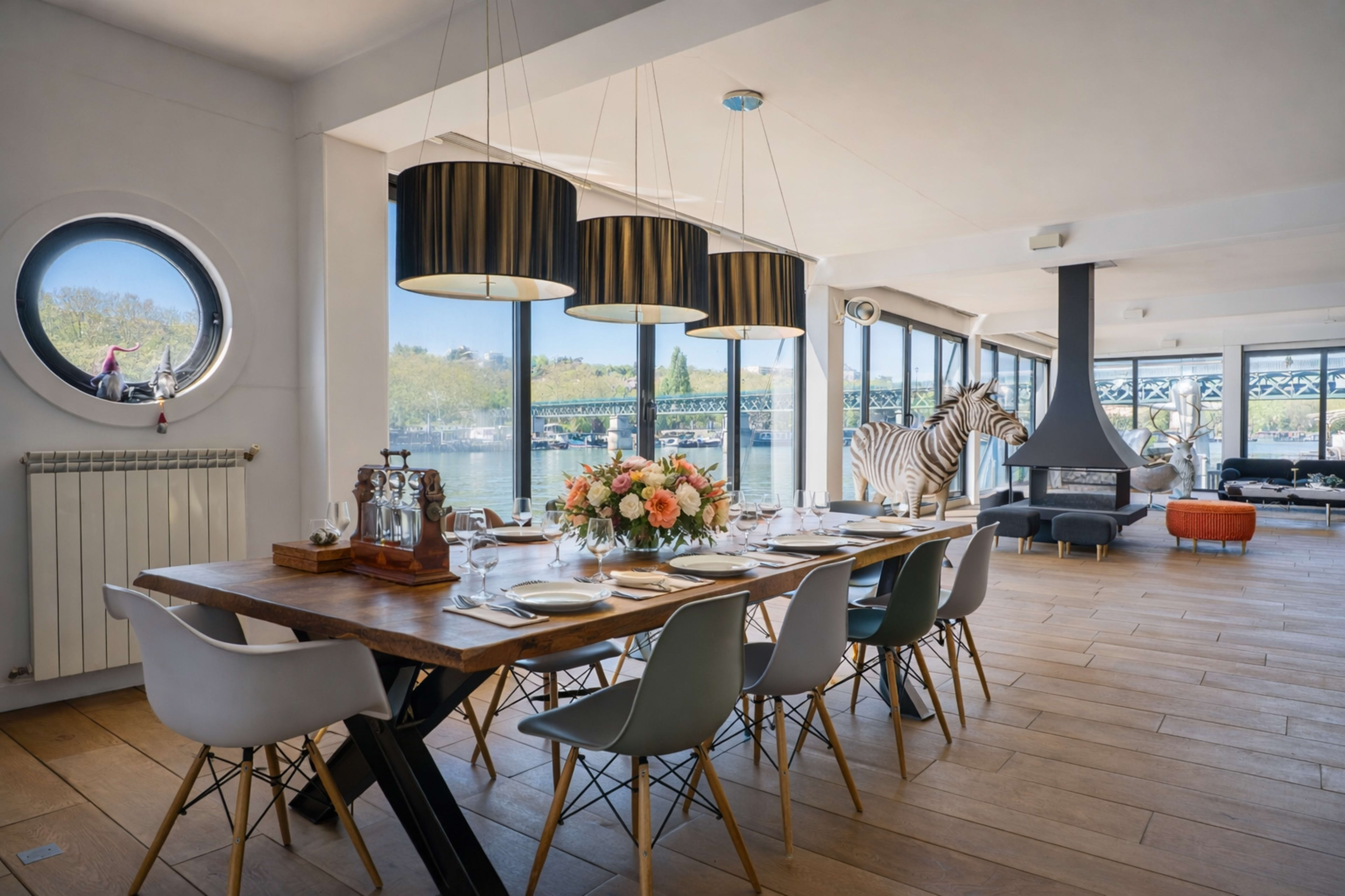 A modern dining area features a long wooden table set for a meal, surrounded by sleek chairs, with large windows offering a view of a river and decorative elements including a zebra statue and stylish pendant lights.