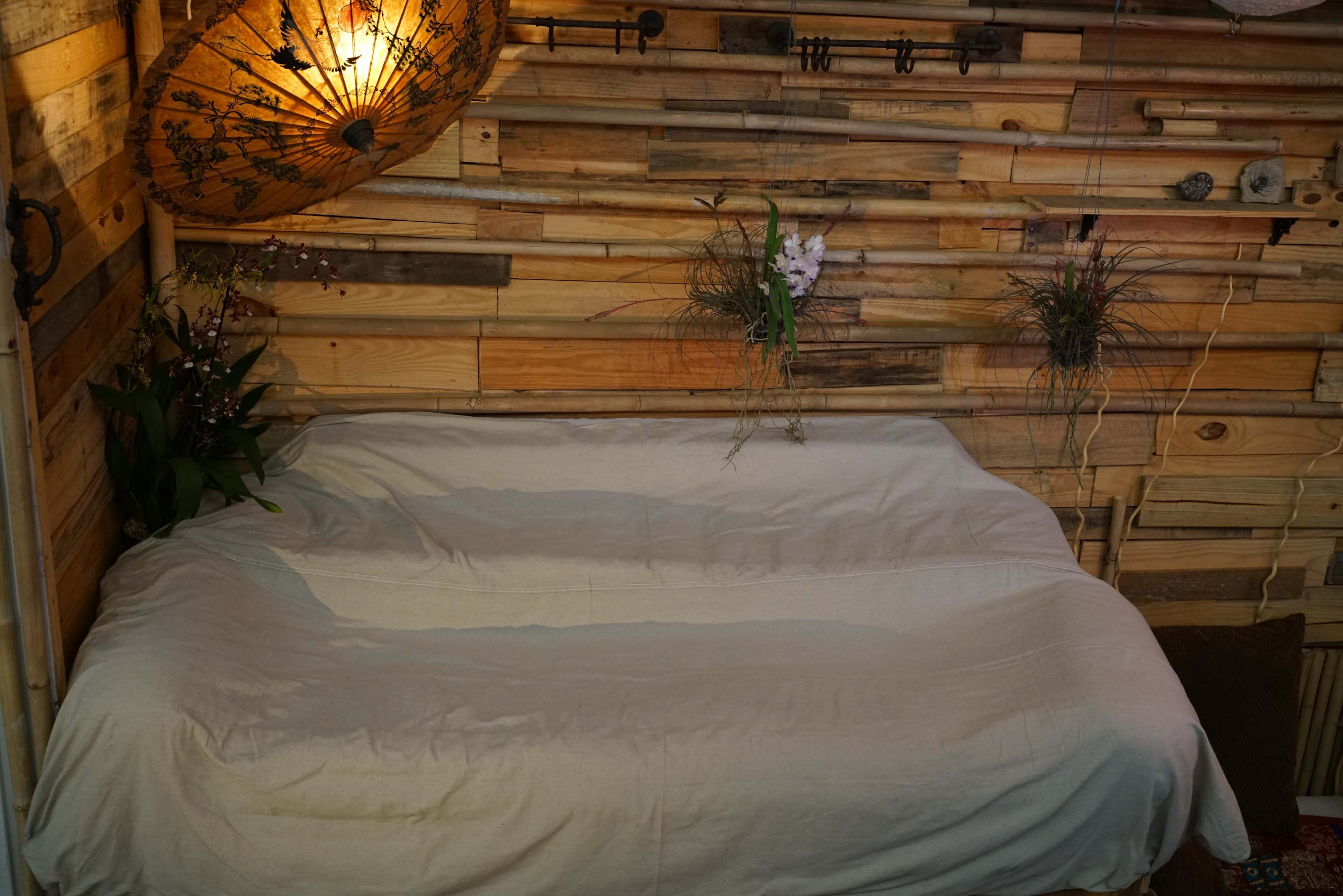 A simple sofa covered with a beige blanket is positioned against a backdrop of wooden planks, accented by hanging plants and a decorative umbrella.