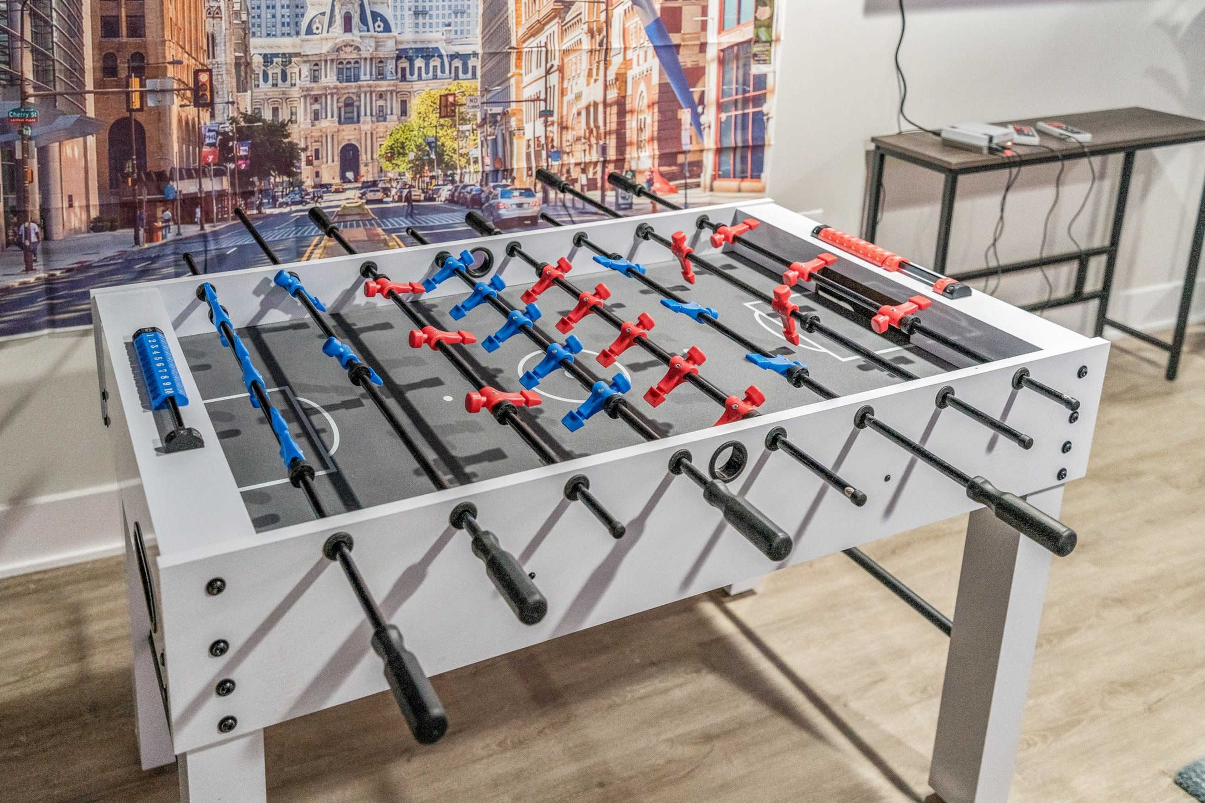 A foosball table with red and blue players is set up in a recreation area, surrounded by urban-themed wall art.