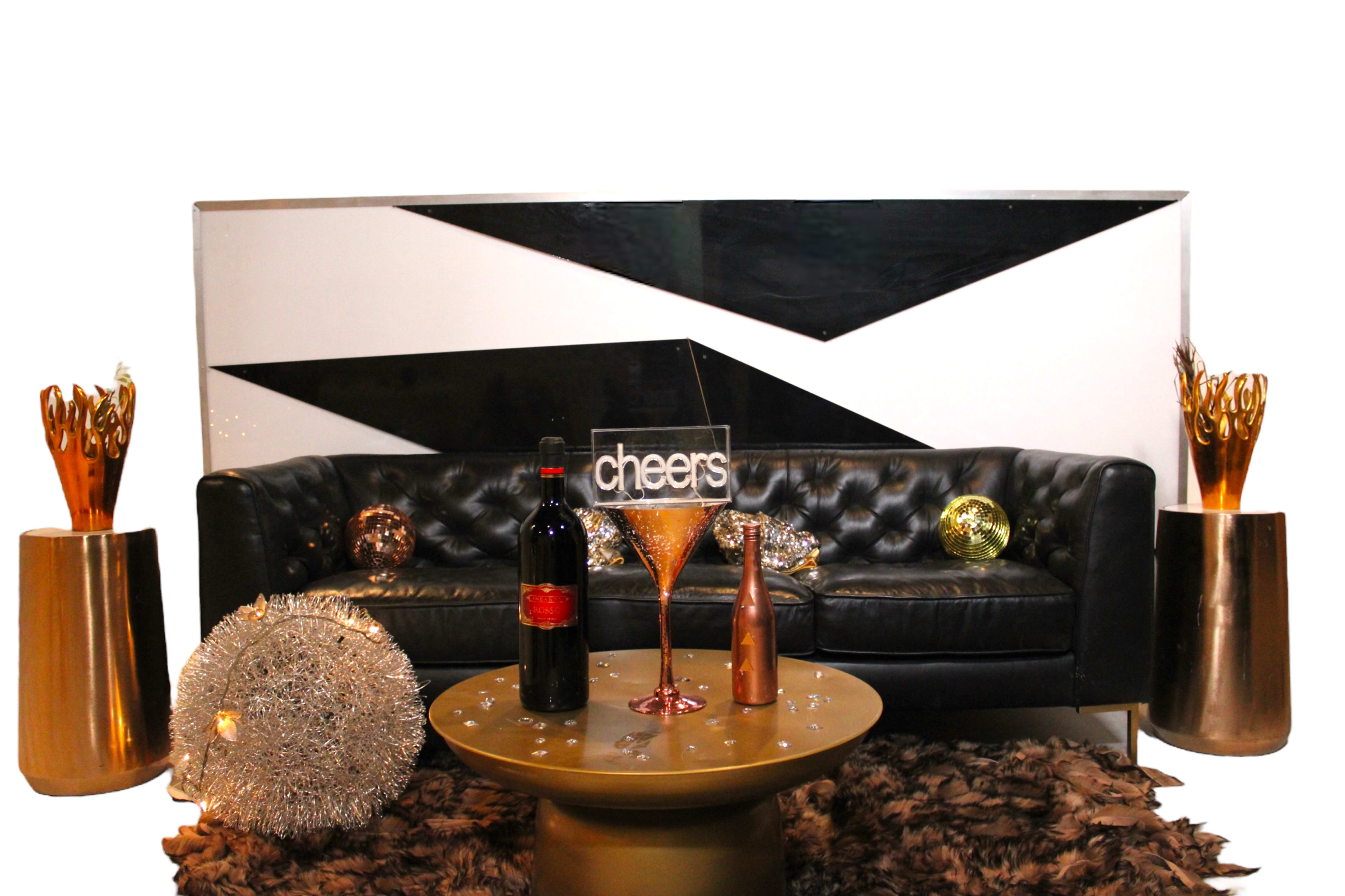 A black couch is placed in front of a modern geometric backdrop, accompanied by decorative gold vases, a round table, and various glasses on the table.