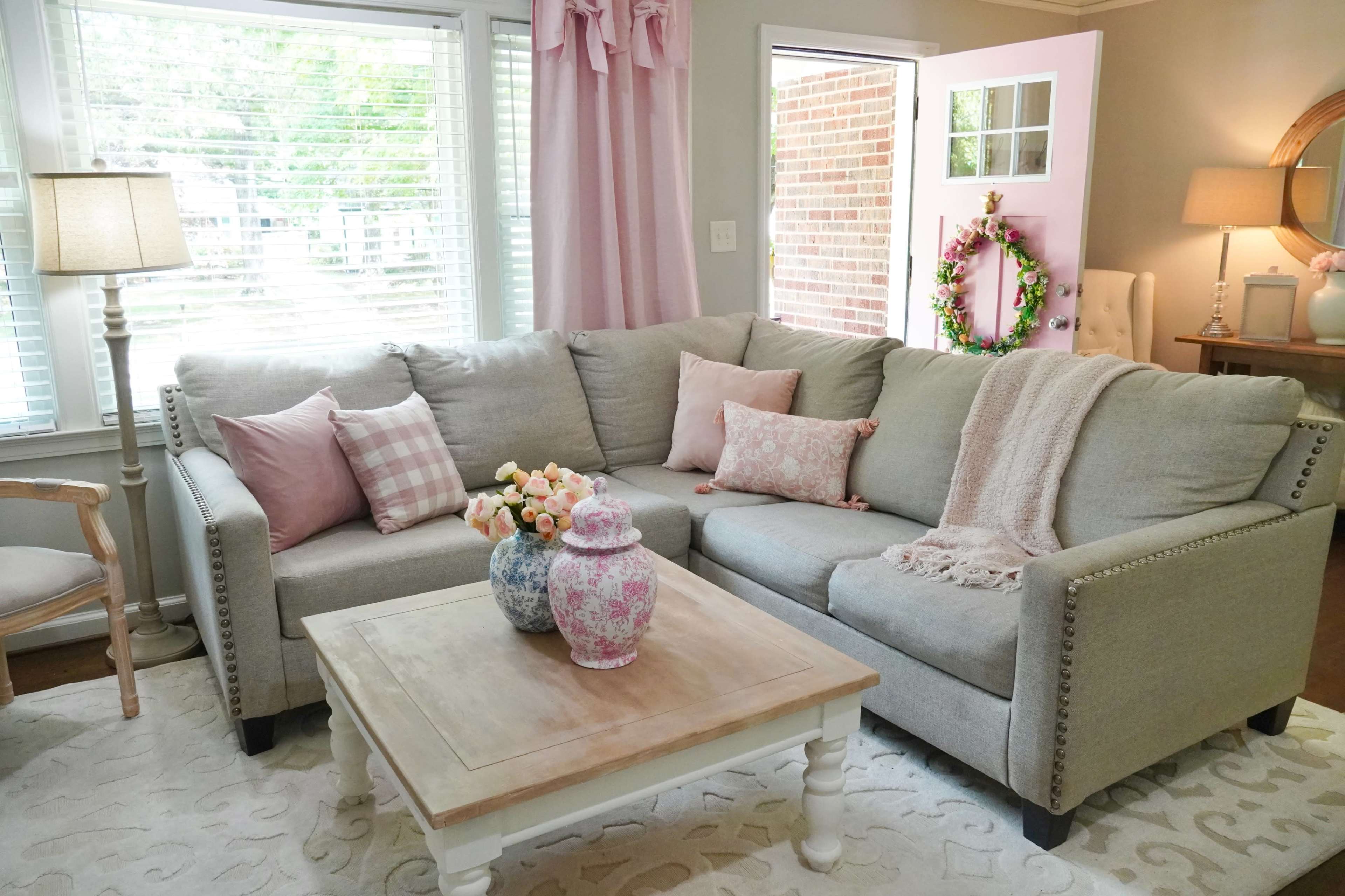 A cozy living room featuring a gray sectional sofa with decorative pillows, a wooden coffee table with floral vases, and large windows allowing natural light.