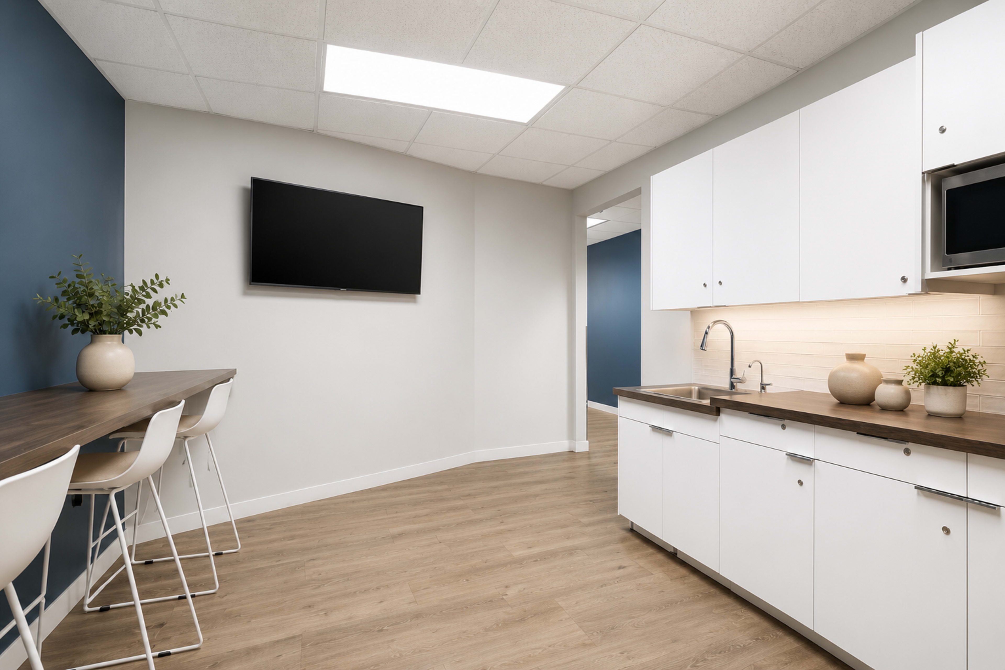 The image shows a modern kitchen area featuring a white cabinetry, a wooden countertop with high stools, and a wall-mounted television.