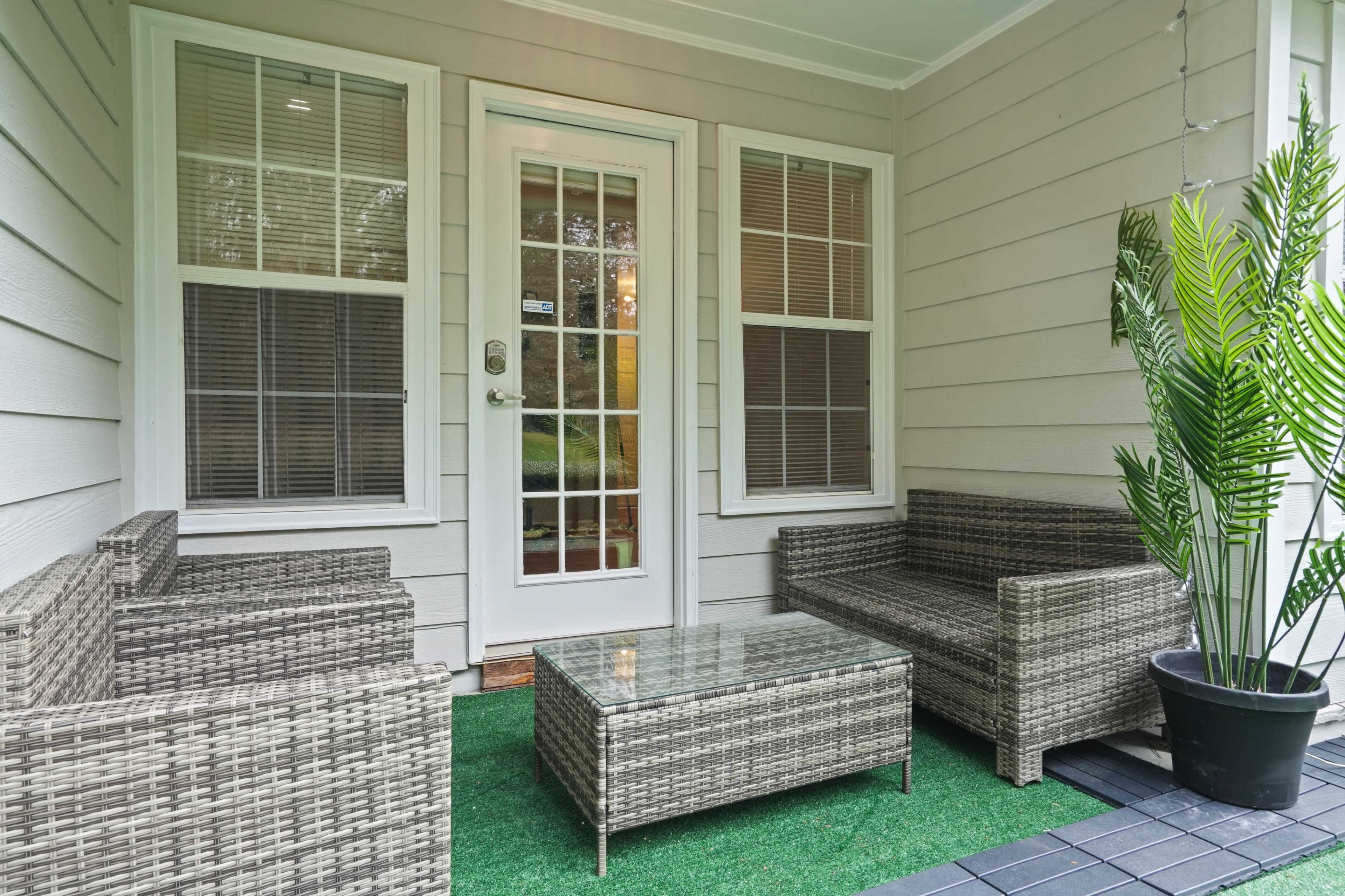 A cozy porch features a glass-topped coffee table and seating made of wicker, surrounded by potted greenery near the entrance.