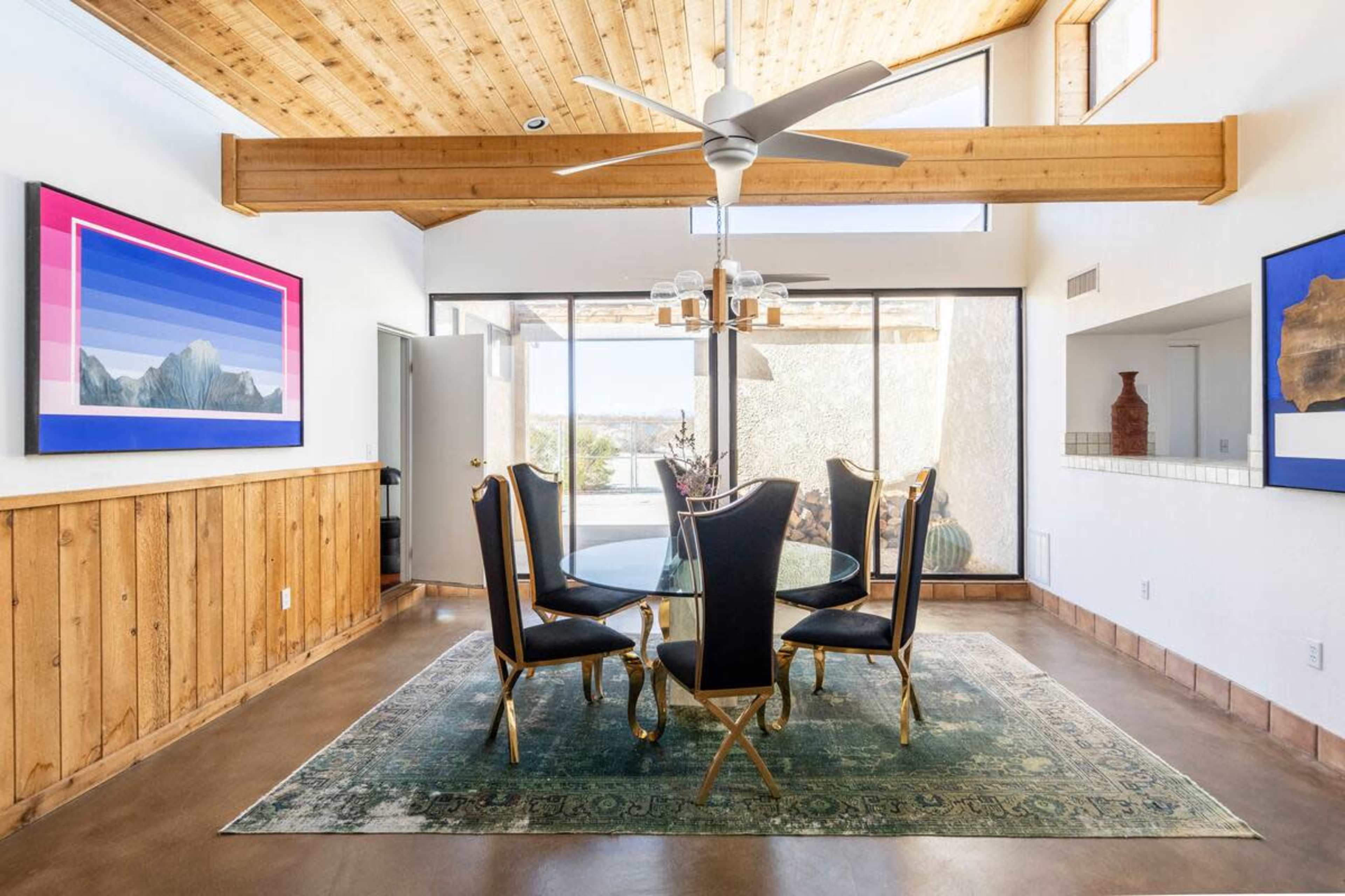 The image shows a dining area with a glass table surrounded by black chairs, set against a backdrop of wooden walls and large windows that provide natural light.