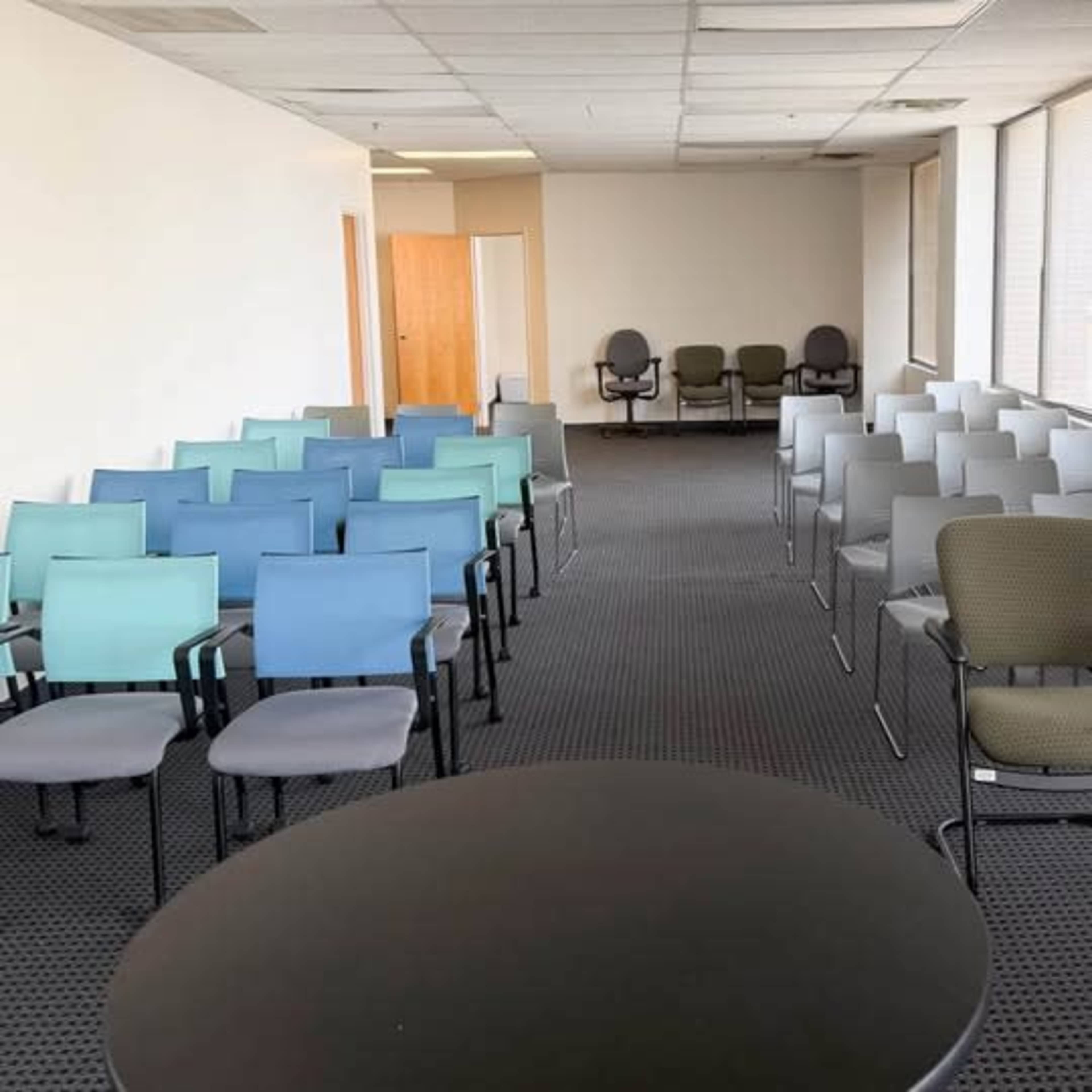 The image shows a large, empty room with rows of multi-colored chairs arranged in two sections, a round black table in the foreground, and a door leading to another area in the background.