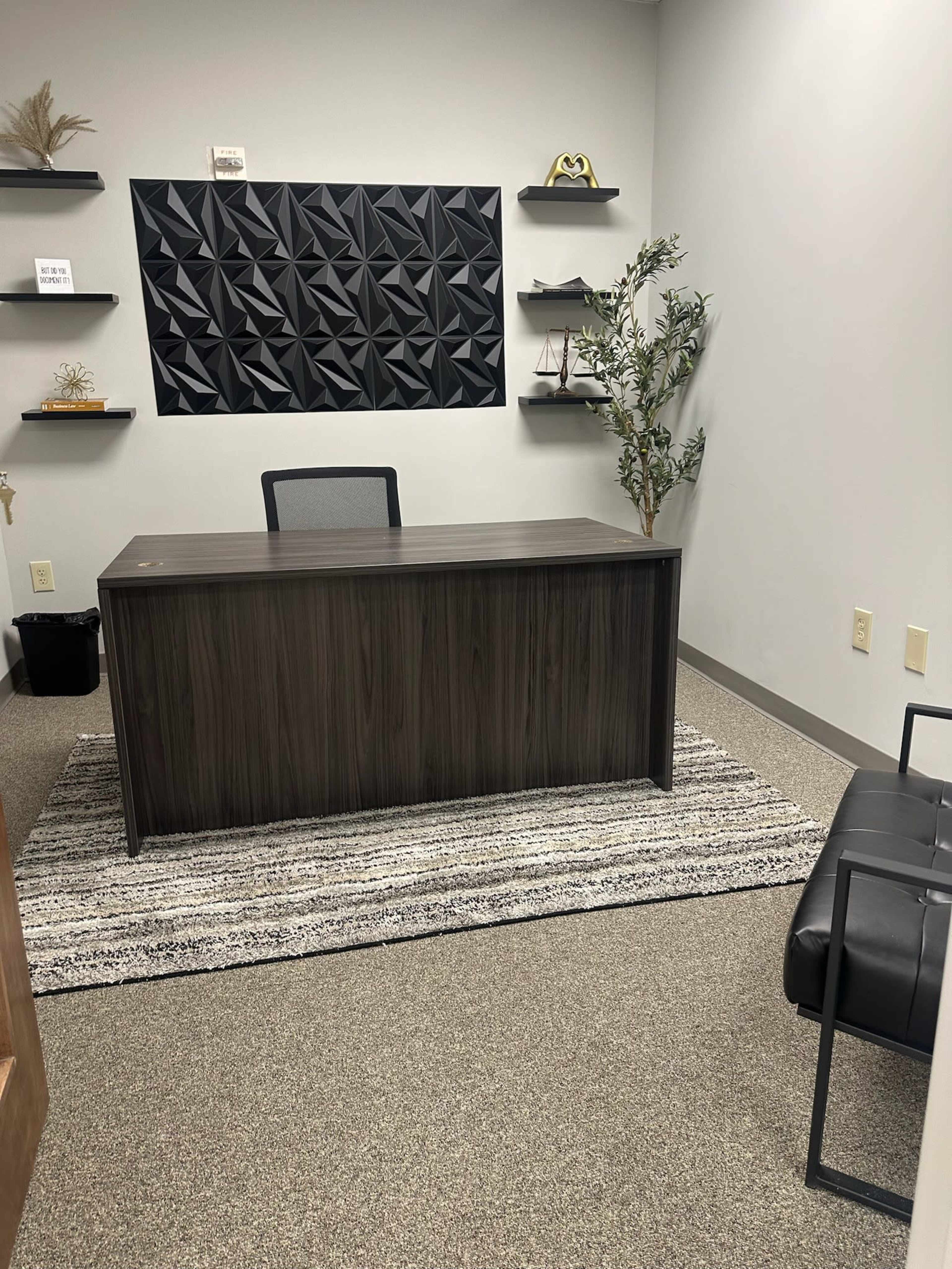 The image shows a modern office space with a dark wooden desk, a chair, a textured wall panel, and shelves displaying decorative items, all on a carpeted floor.