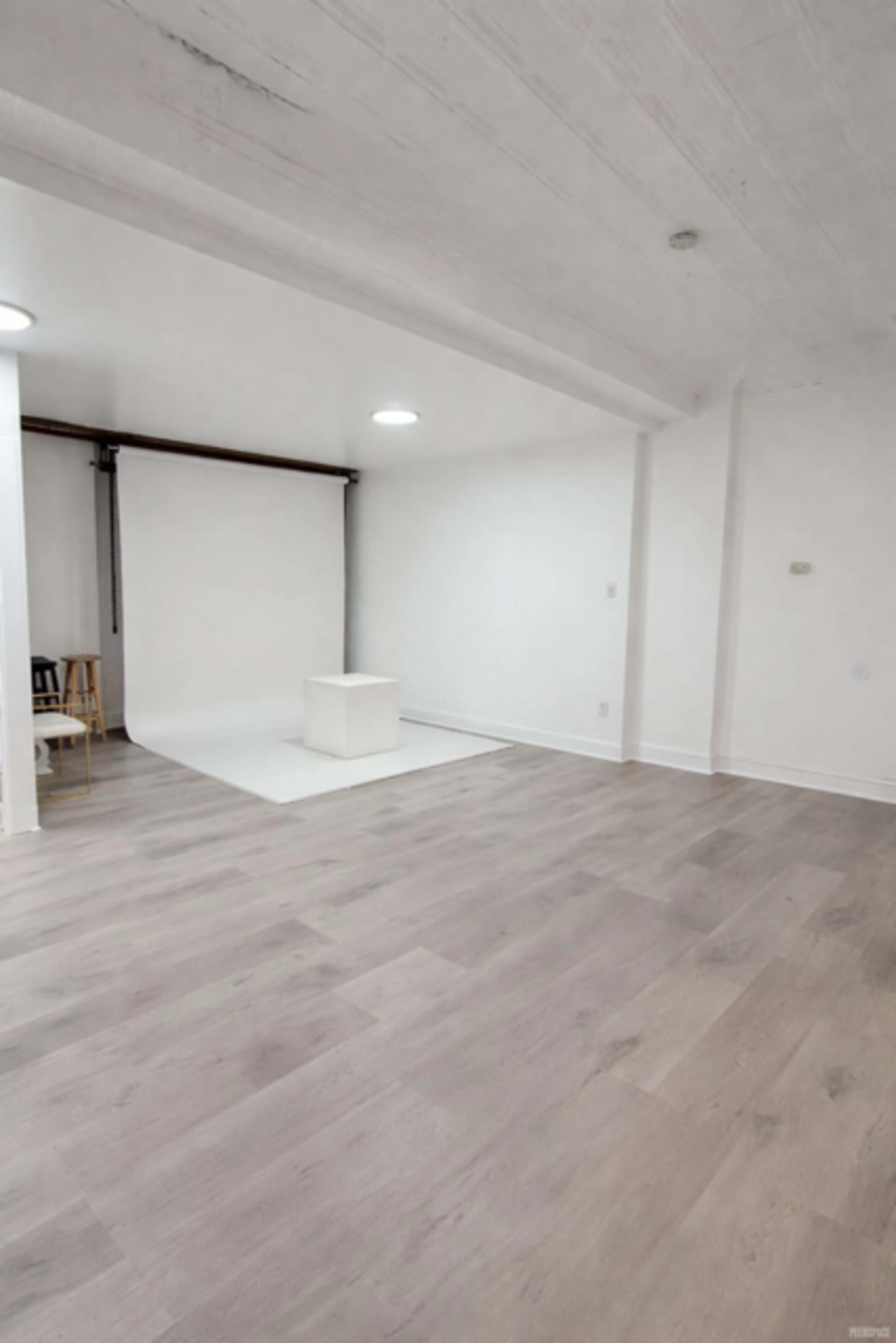 The image shows a spacious, minimalist interior with white walls, light wooden flooring, and a blank studio backdrop in the corner.