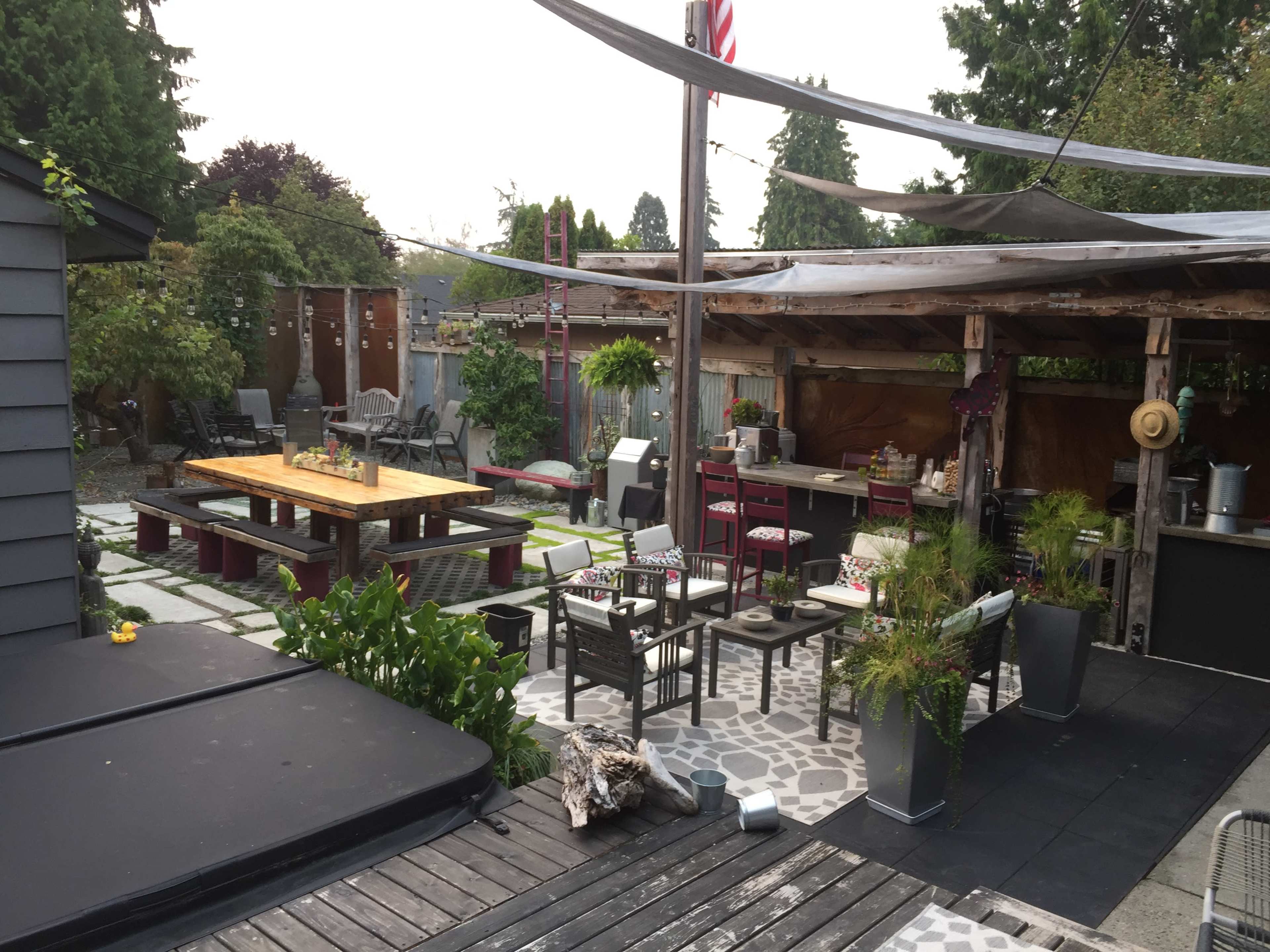 A backyard patio with a large wooden dining table, various seating areas, and a covered outdoor kitchen among landscaped greenery.