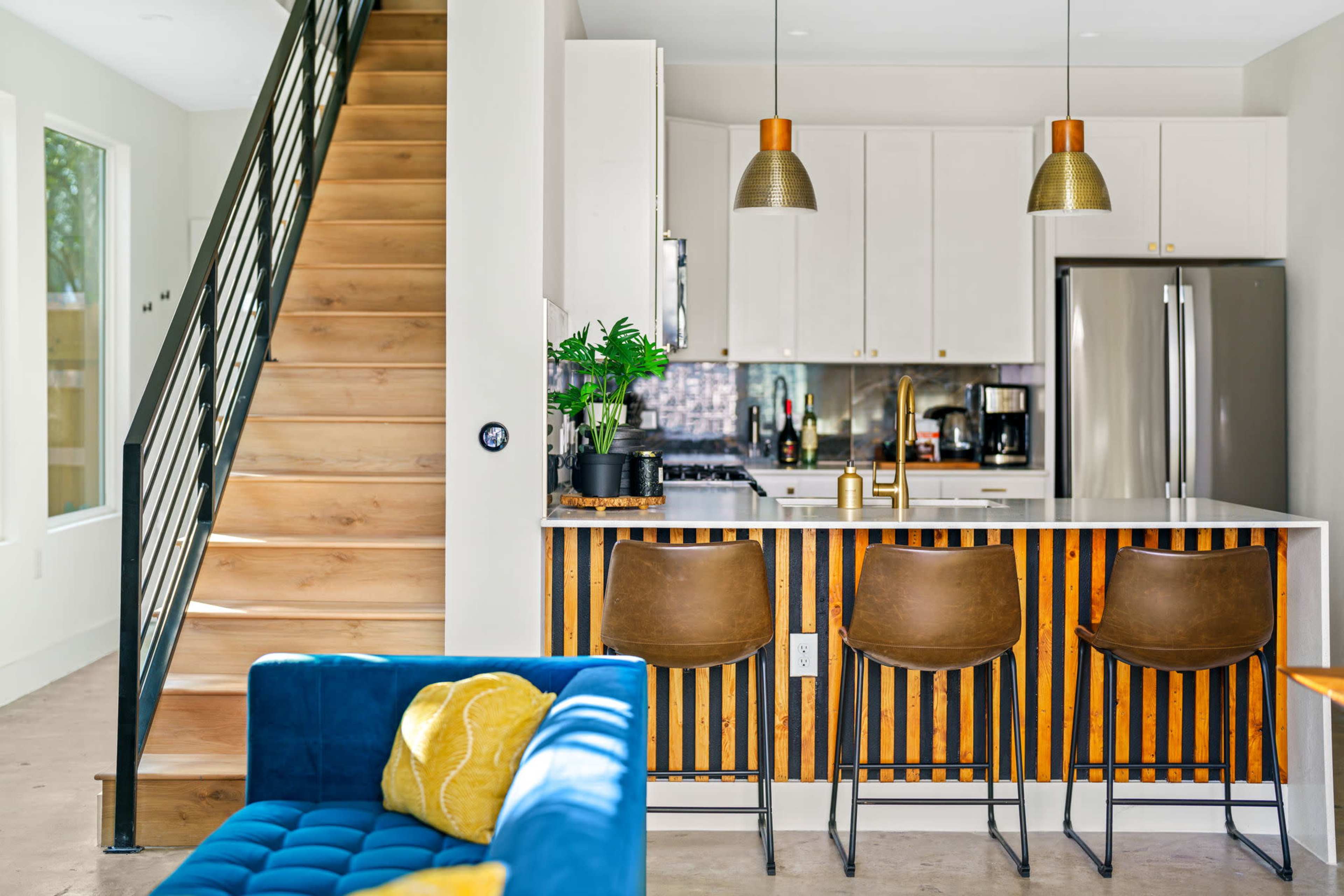 A modern kitchen area with a stairway to the left, featuring a bar with three stools and a countertop adorned with plants and kitchen appliances.