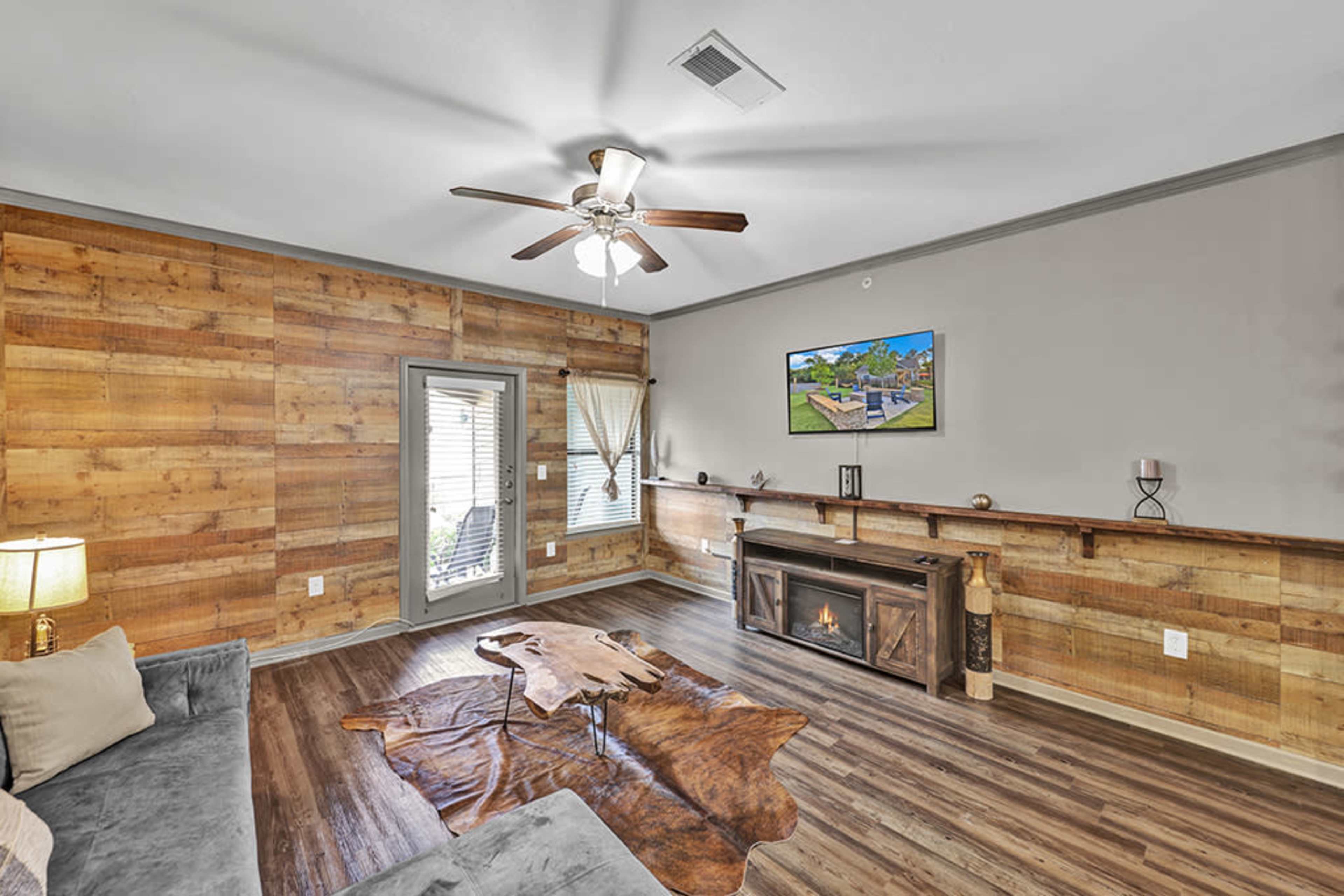 The living room features wooden walls, a ceiling fan, a TV mounted above a decorative fireplace, and a large area rug.
