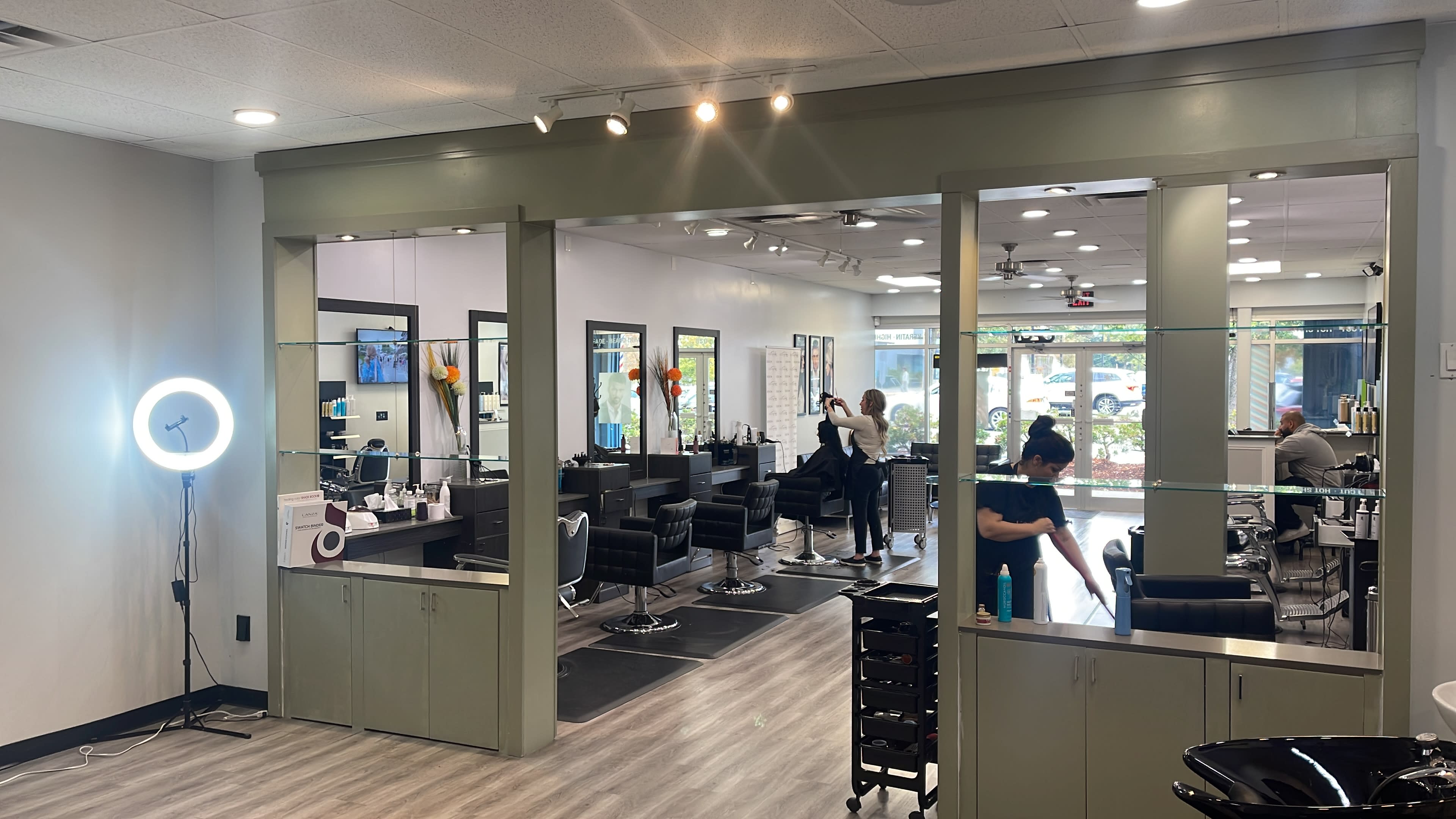 The image shows the interior of a hair salon with black styling chairs, mirrors, and a reception area, while stylists attend to clients.