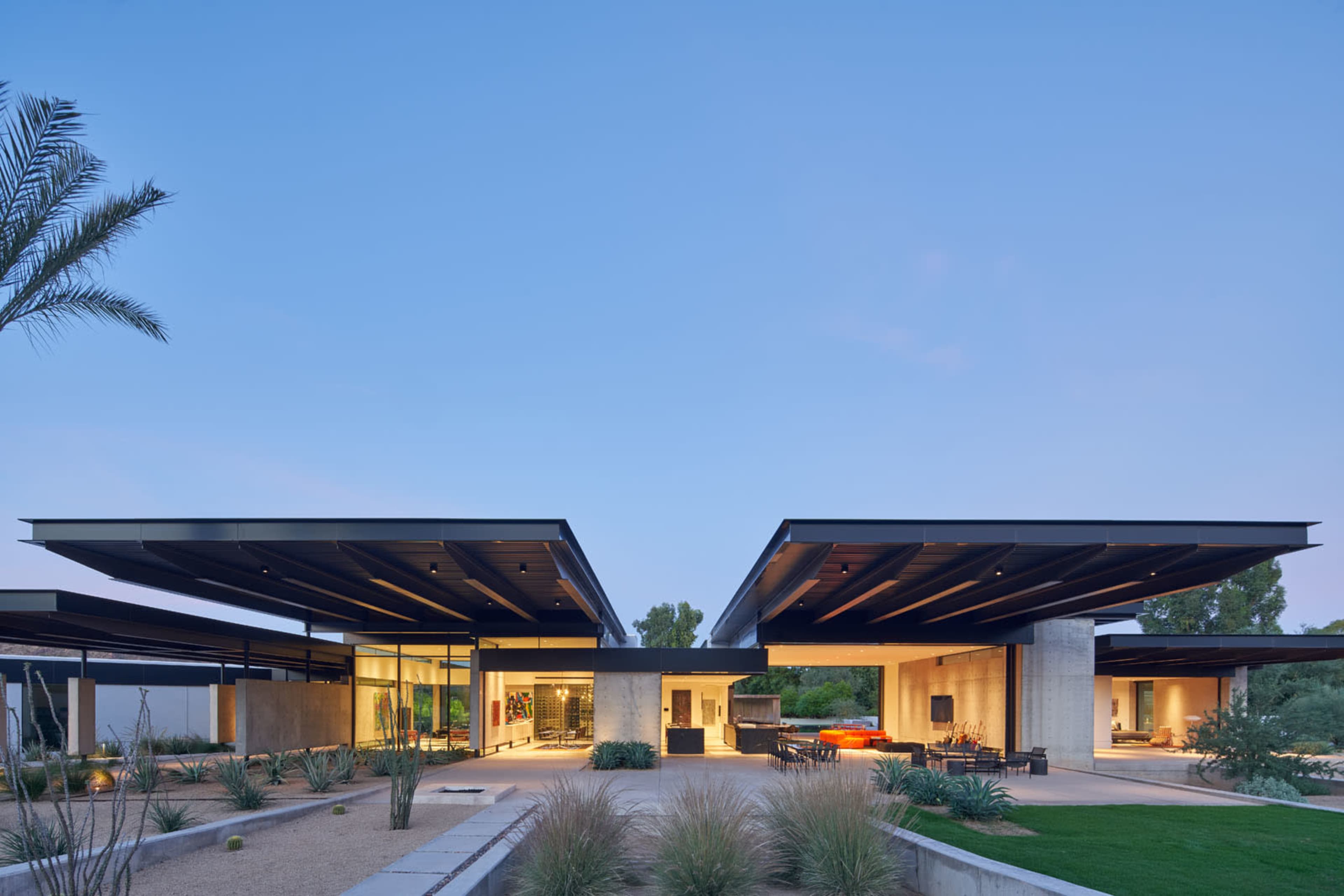 A modern house features an open design with a large central area and cantilevered roofs, surrounded by desert landscaping.