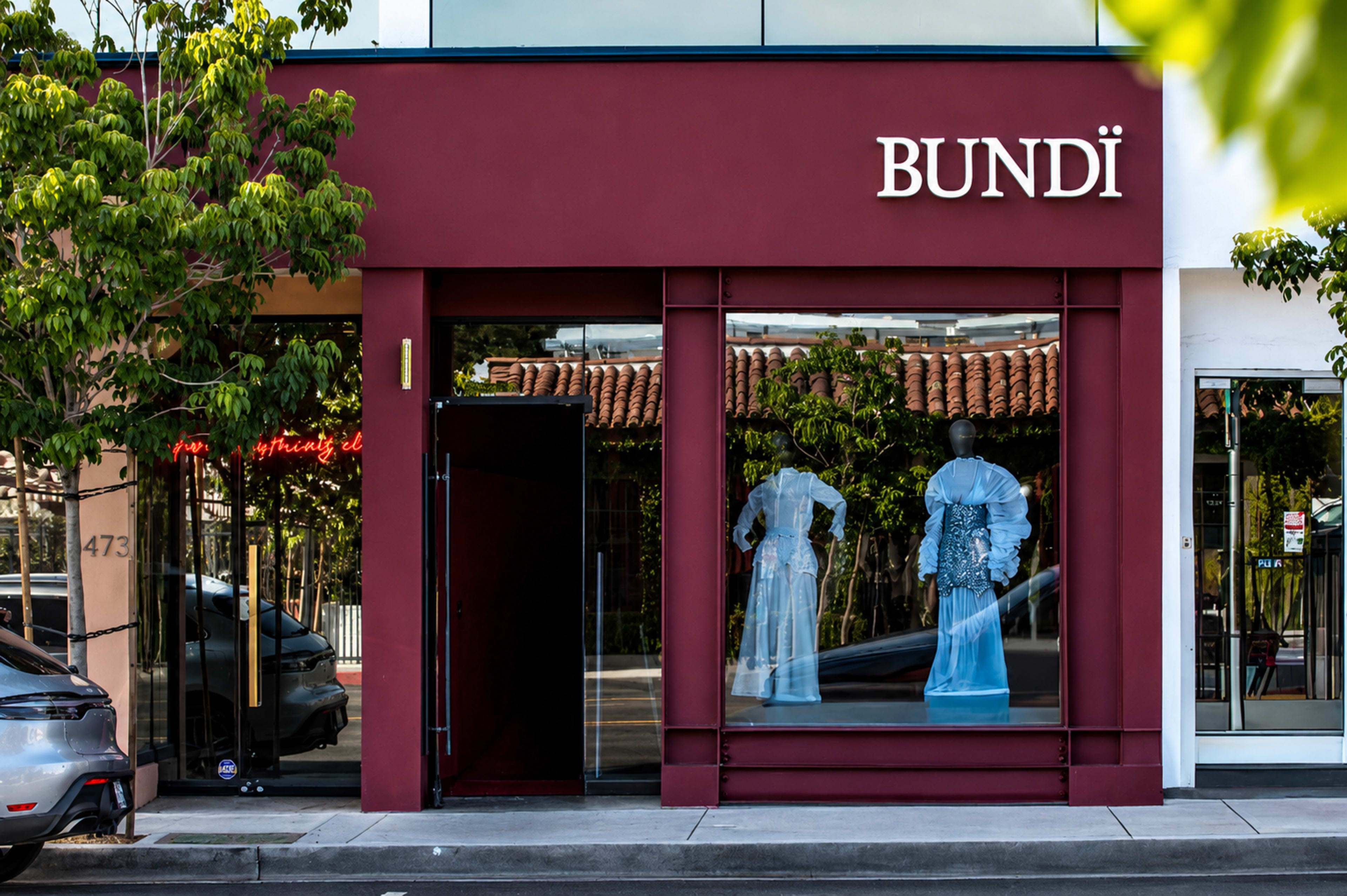The storefront of a boutique named "BUNDÏ," featuring mannequins dressed in blue clothing, is displayed behind large glass windows surrounded by greenery.