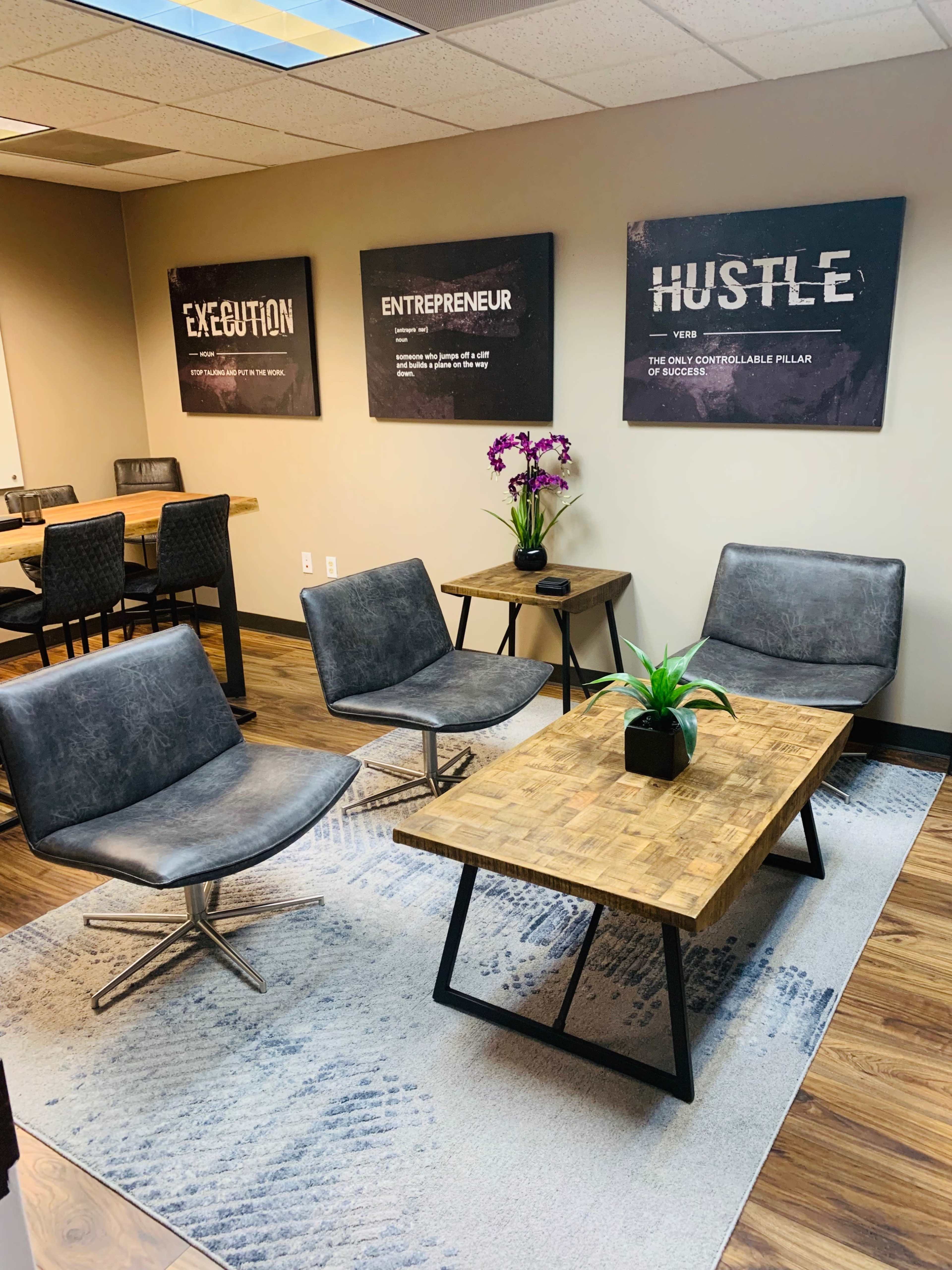 A modern office waiting area features four gray chairs arranged around a wooden coffee table, with motivational wall art and a potted plant.