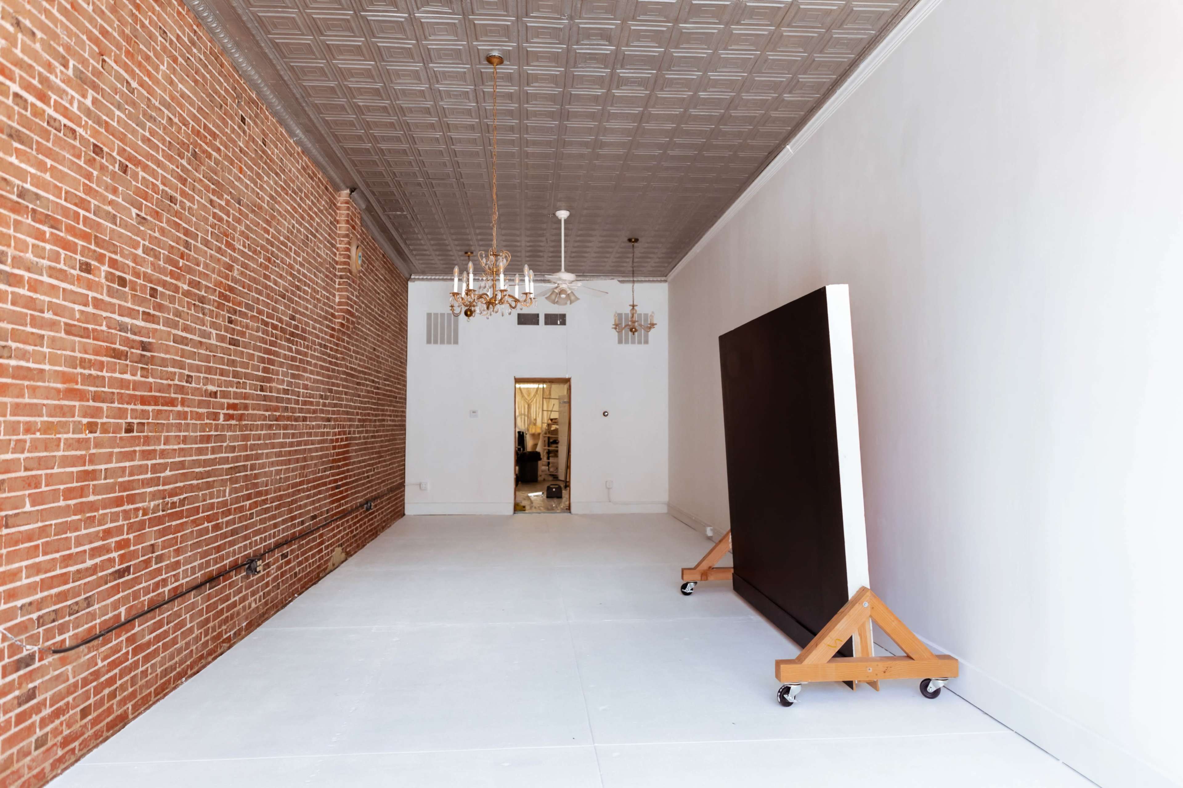 The image shows a spacious, empty room with exposed brick walls, a white ceiling, and a large piece of art on a wooden easel near the center.