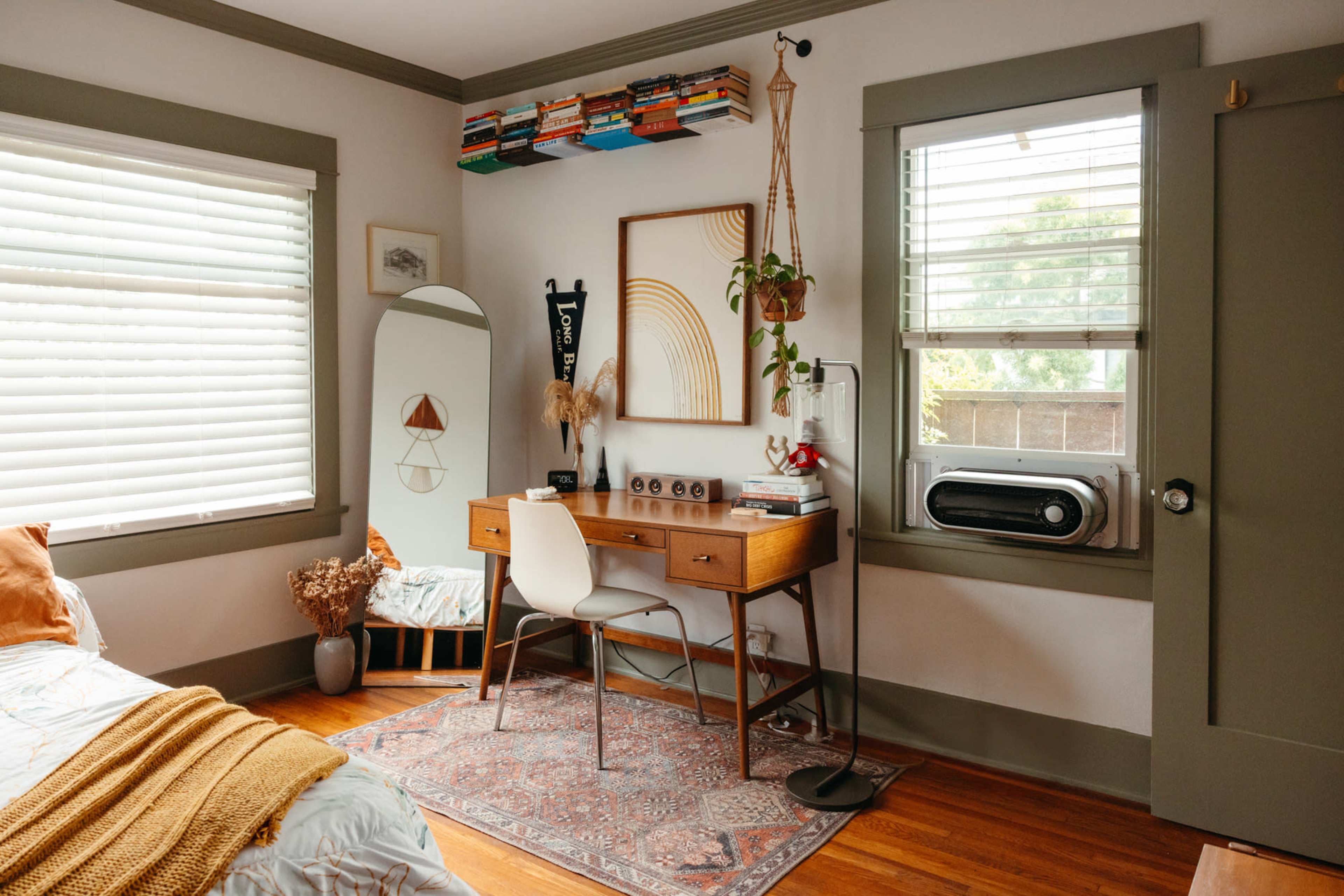 A cozy bedroom with a wooden desk, a chair, a large mirror, and shelves displaying books above the window.