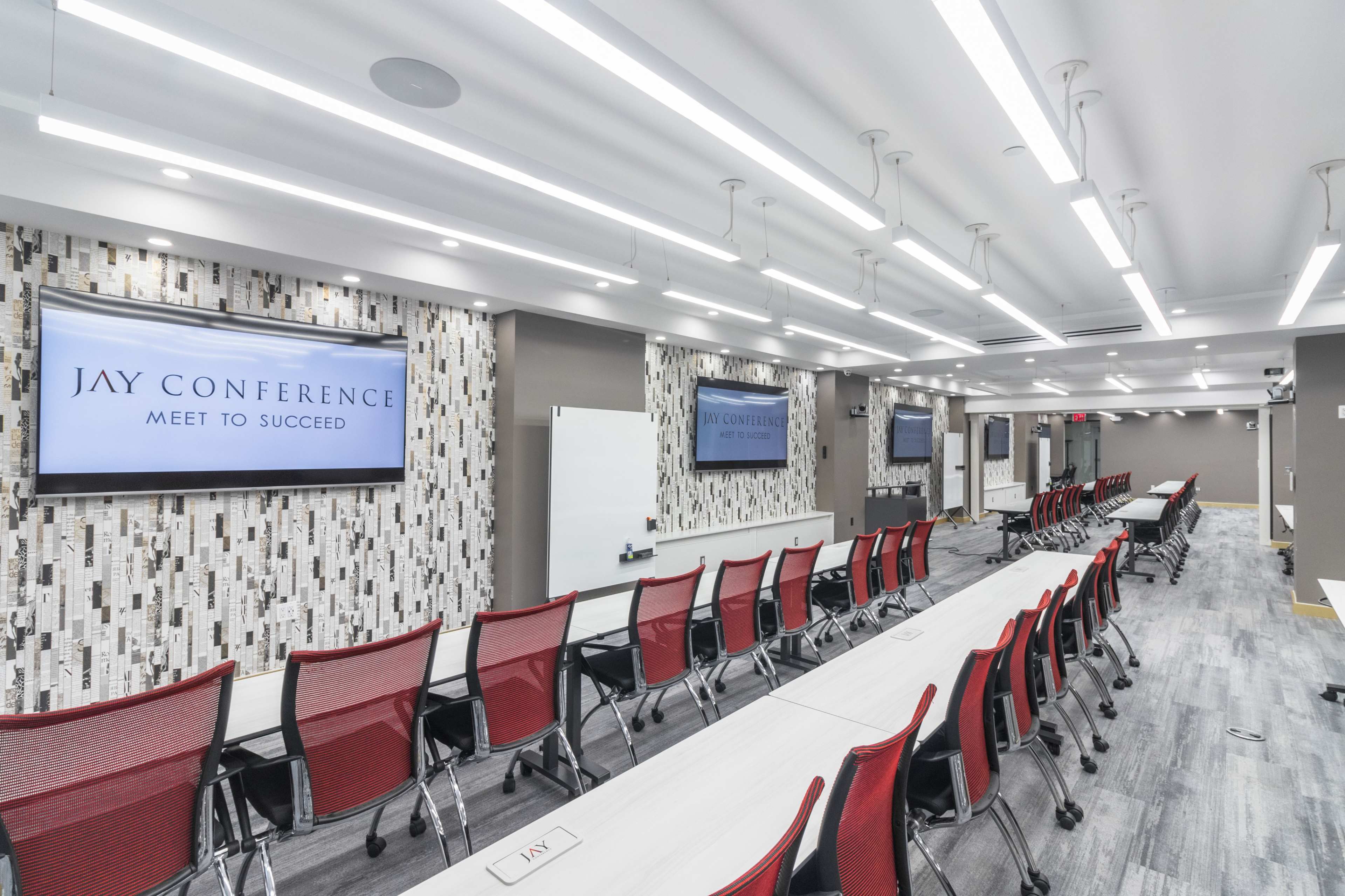 The image shows a modern conference room with red chairs arranged around long tables and two large screens displaying "JAY CONFERENCE."