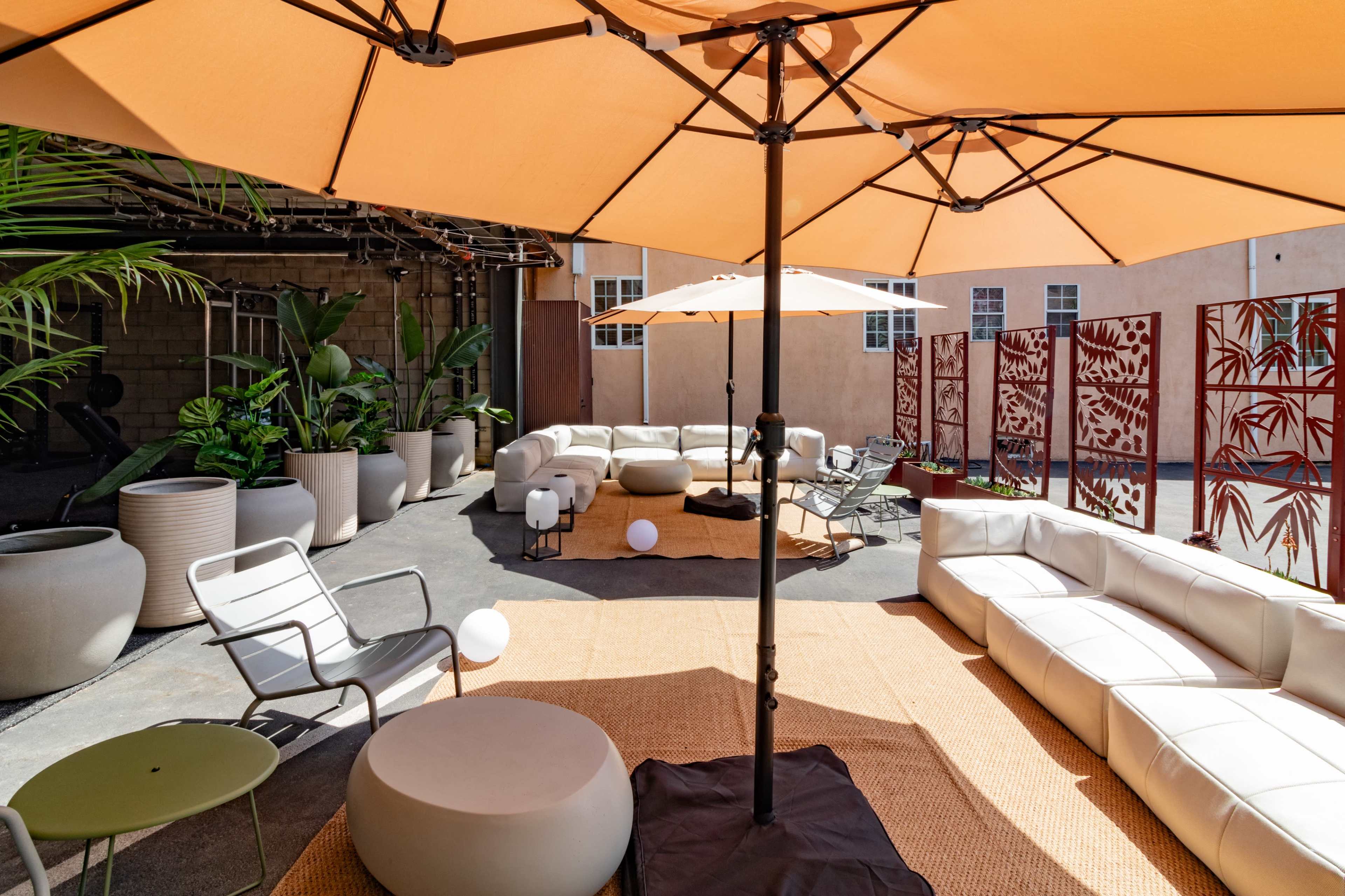 A modern outdoor lounge area featuring white sectional sofas, large umbrellas, potted plants, and a decorative screen.