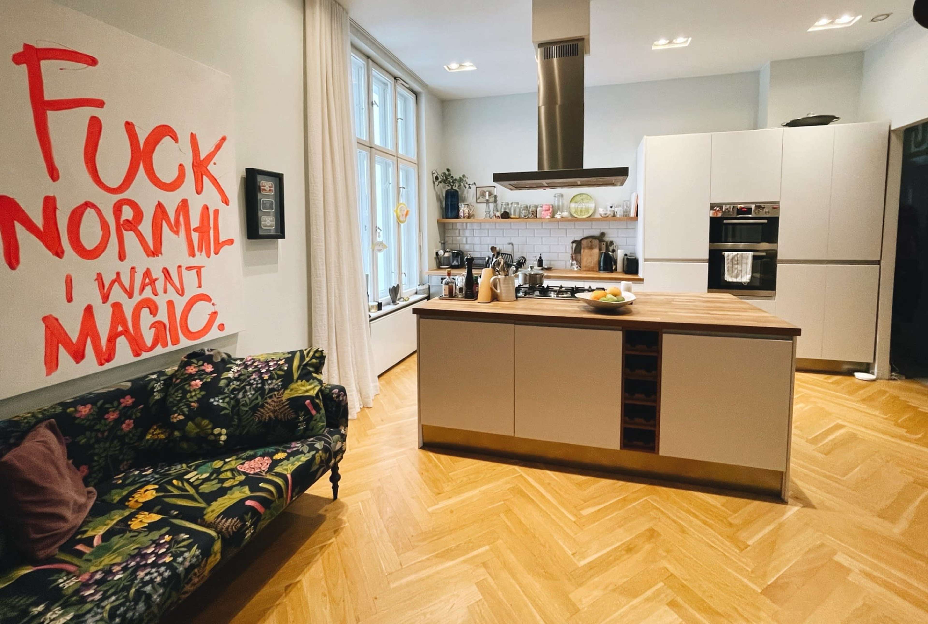 A modern kitchen with a central island, floral-patterned sofa, and a large wall art piece with the words "FUCK NORMAL I WANT MAGIC."