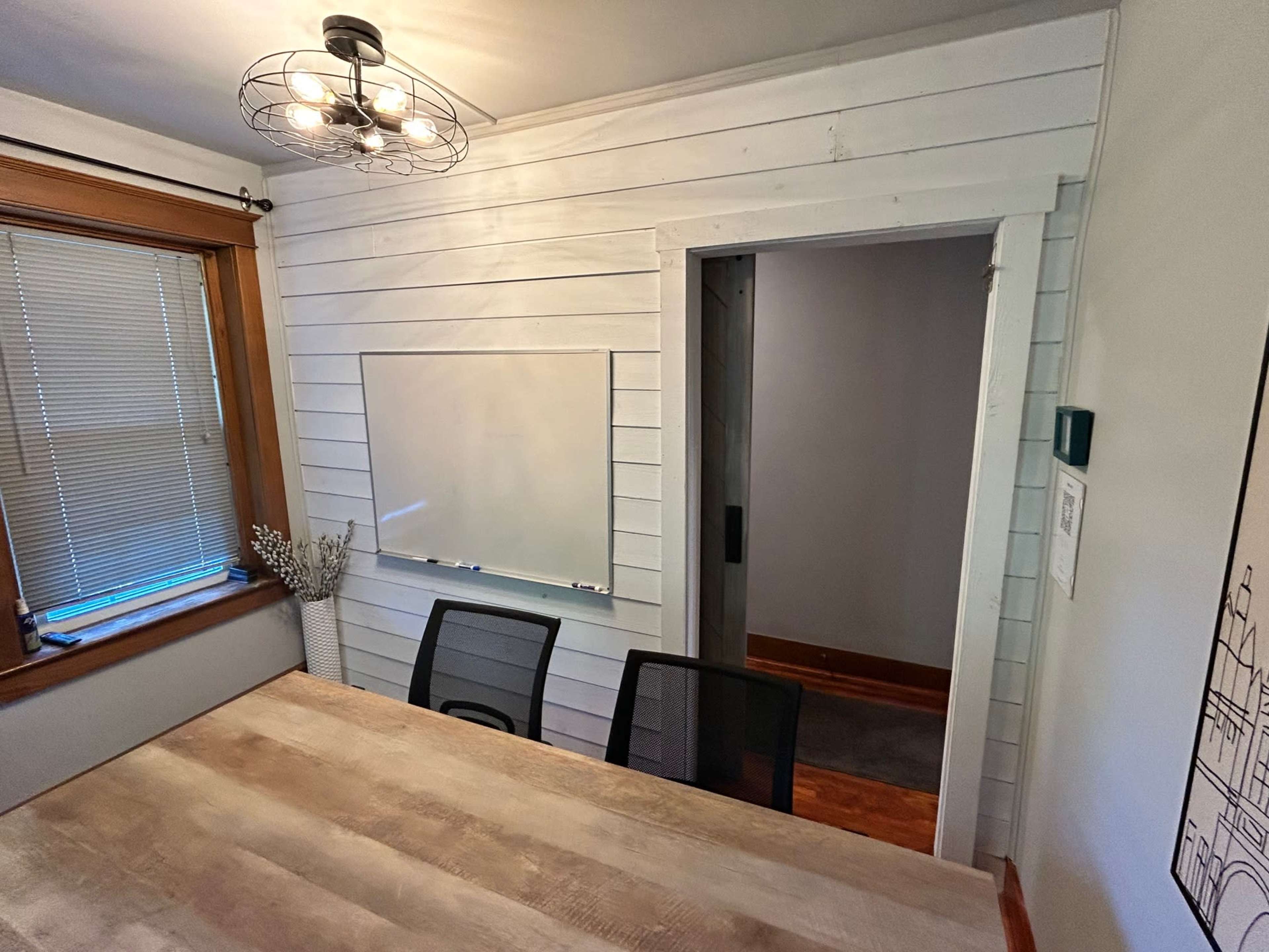 The image shows a well-lit room with a wooden table, three black chairs, a whiteboard mounted on the wall, and a doorway leading to another space.