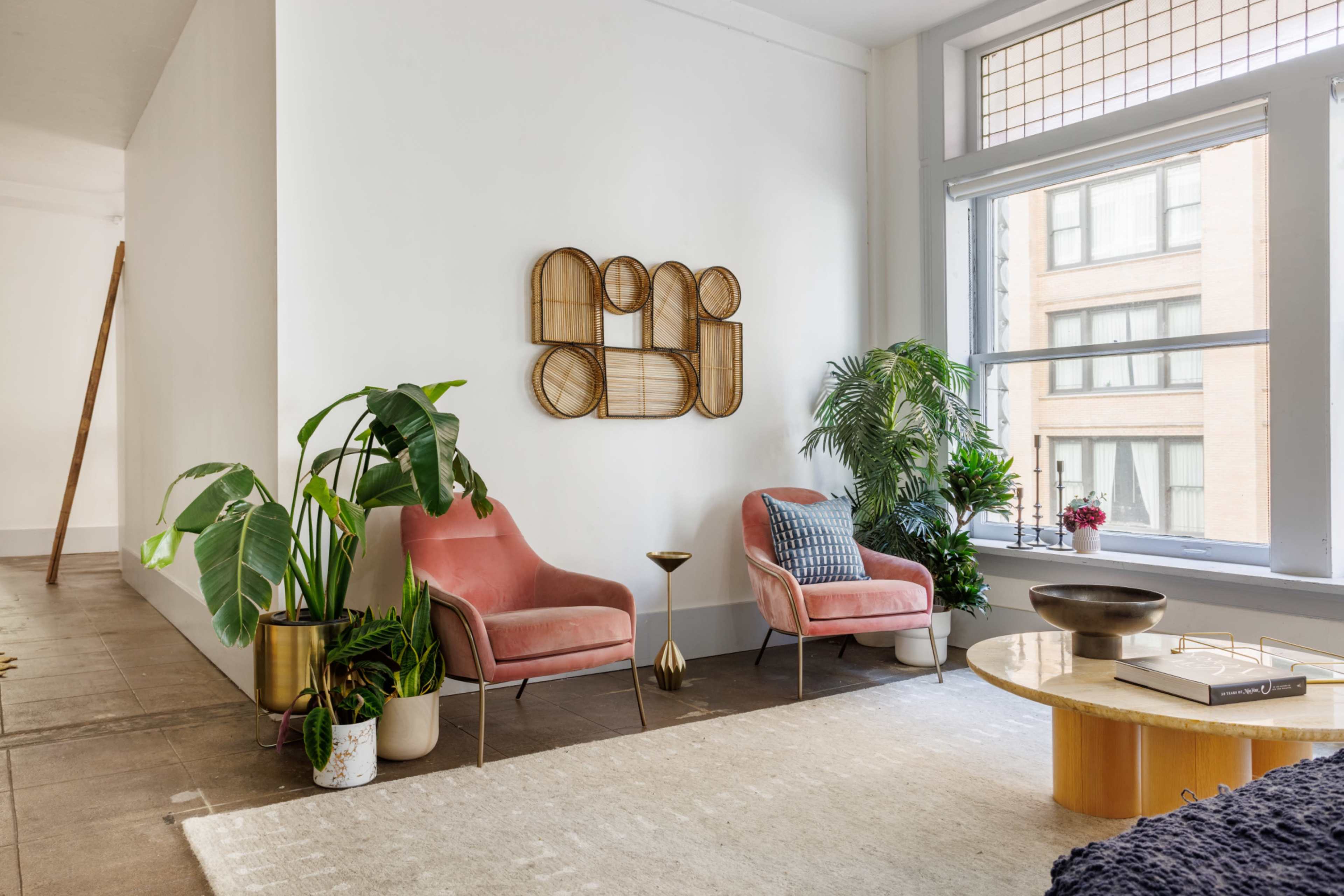 A bright living space features two pink chairs, a round wooden coffee table, and various plants near a large window.