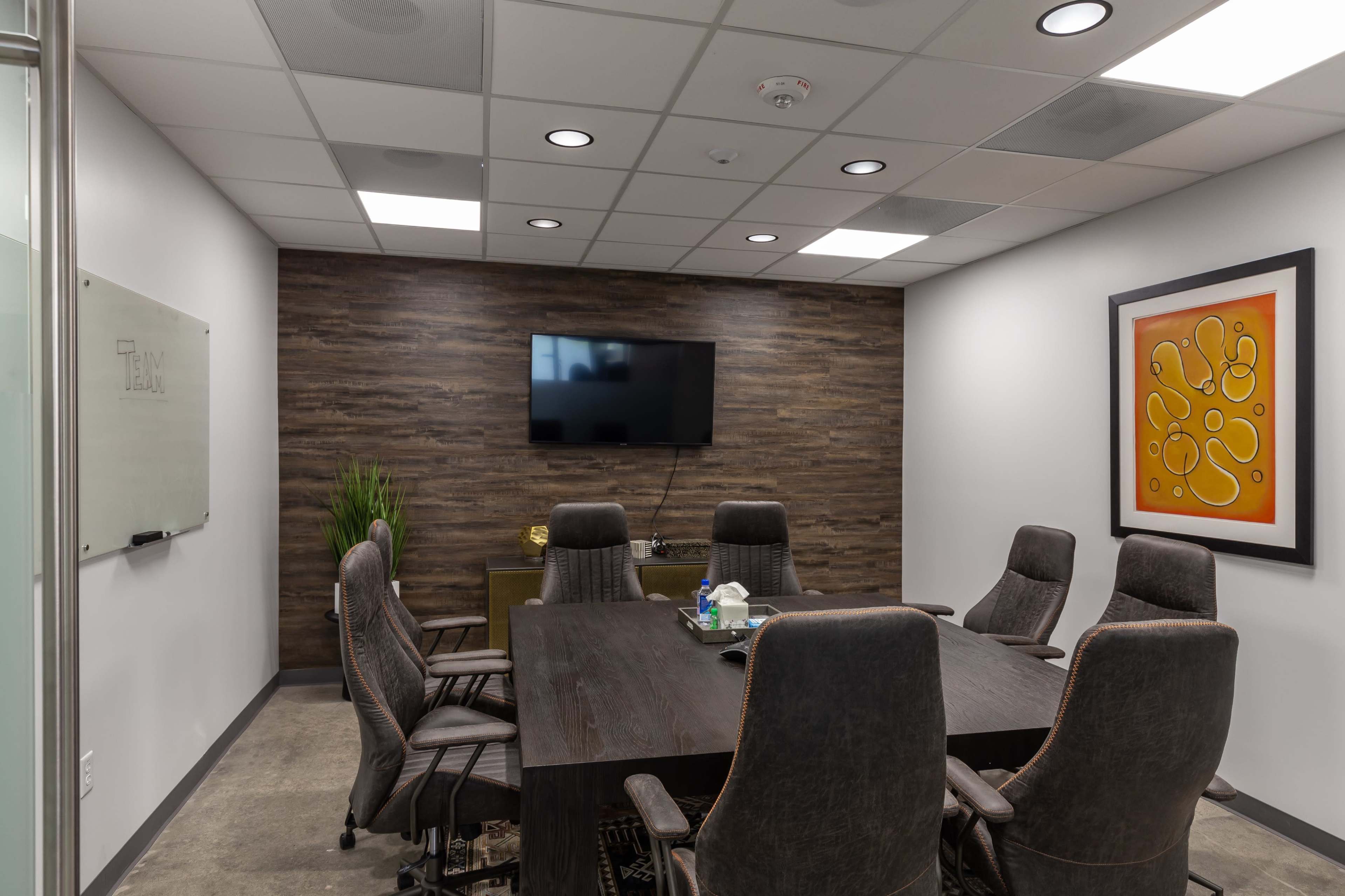 A modern conference room featuring a large wooden table surrounded by eight upholstered chairs, a wall-mounted television, and a whiteboard.