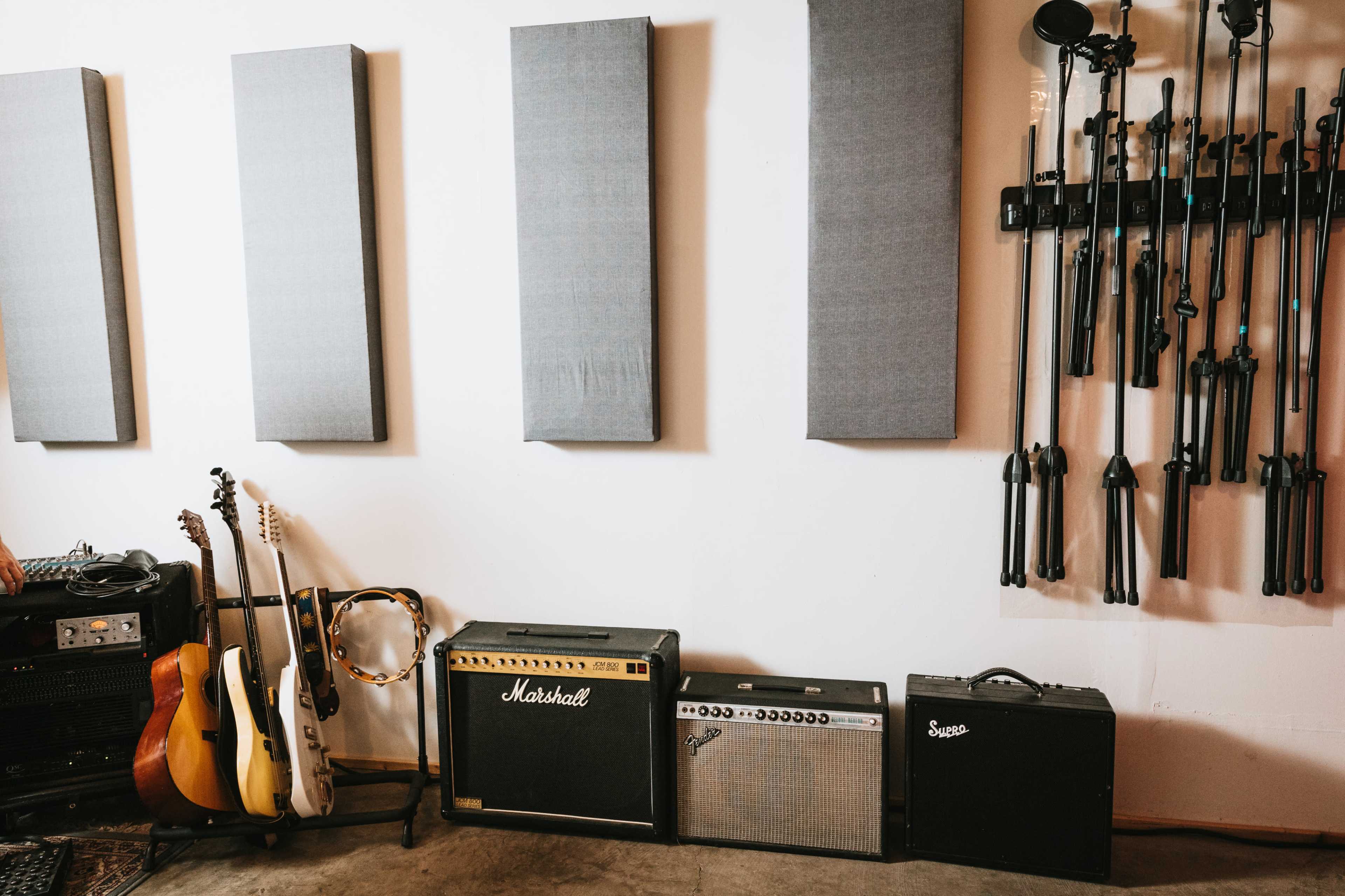 An indoor space with guitars resting against the wall, amplifiers on the floor, and soundproof panels mounted vertically on the wall alongside microphone stands.