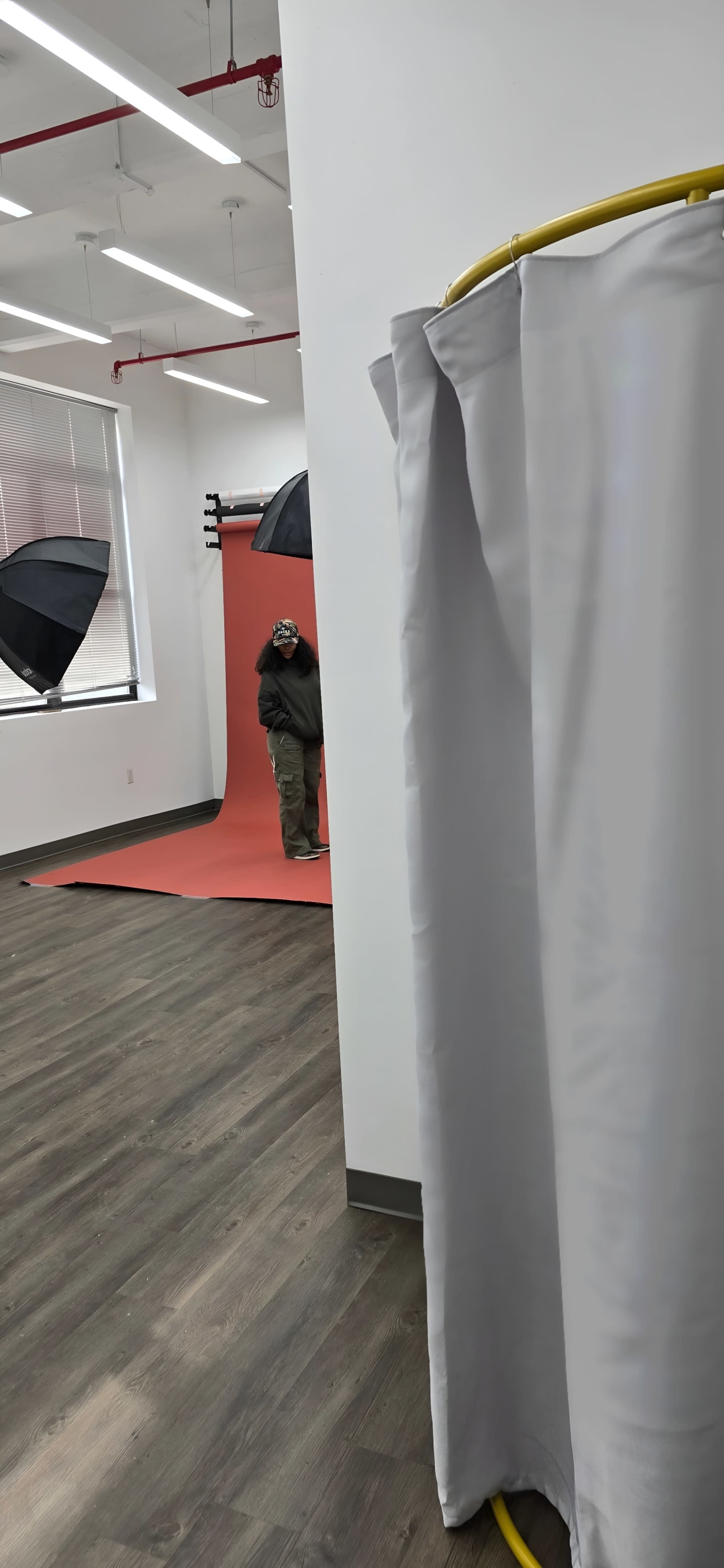 A person is partially visible behind a curtain in a photography studio, with a red backdrop and several umbrellas set up for lighting.
