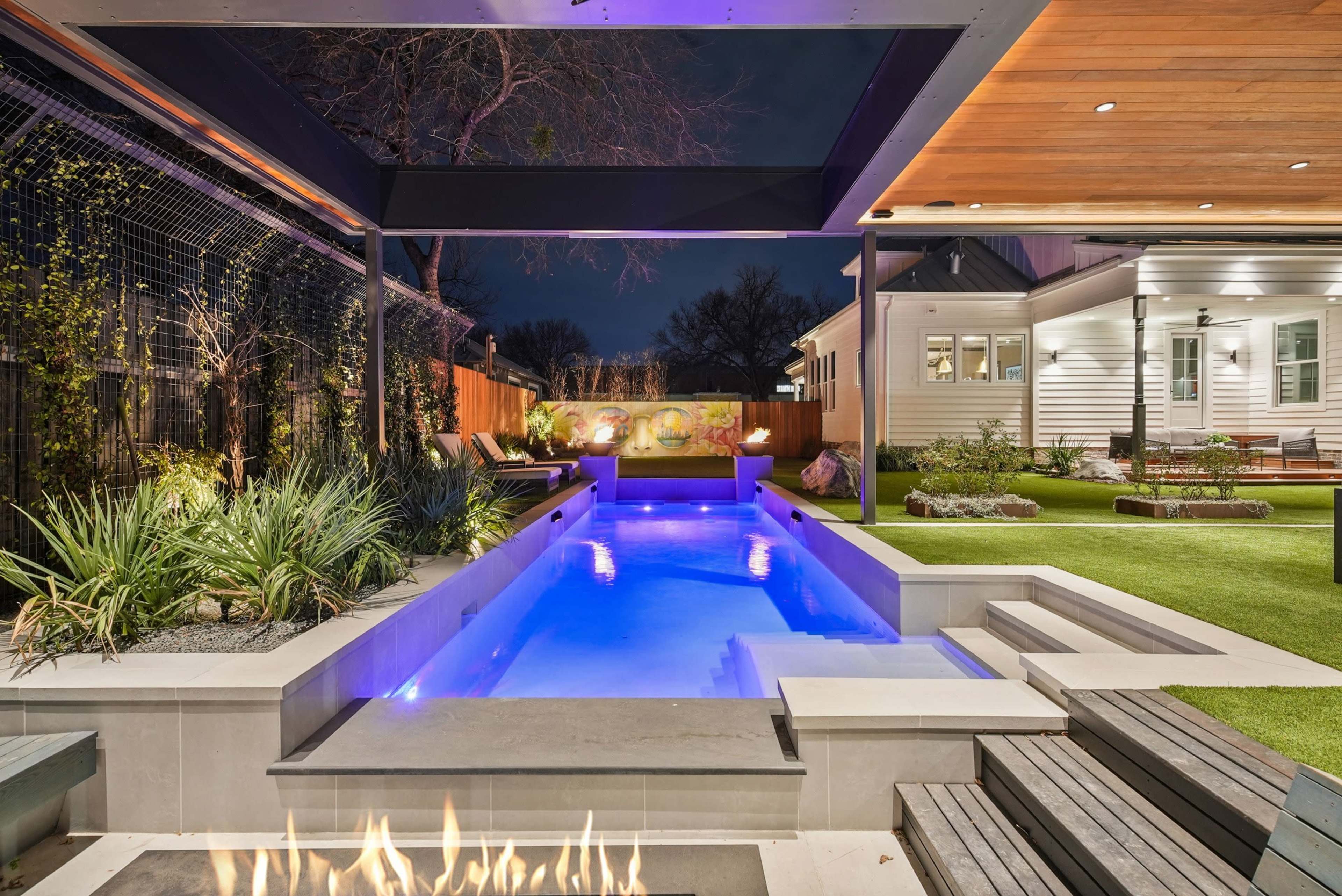 The image shows a modern backyard with a rectangular pool, a fire pit, and landscaped areas illuminated by night lighting.