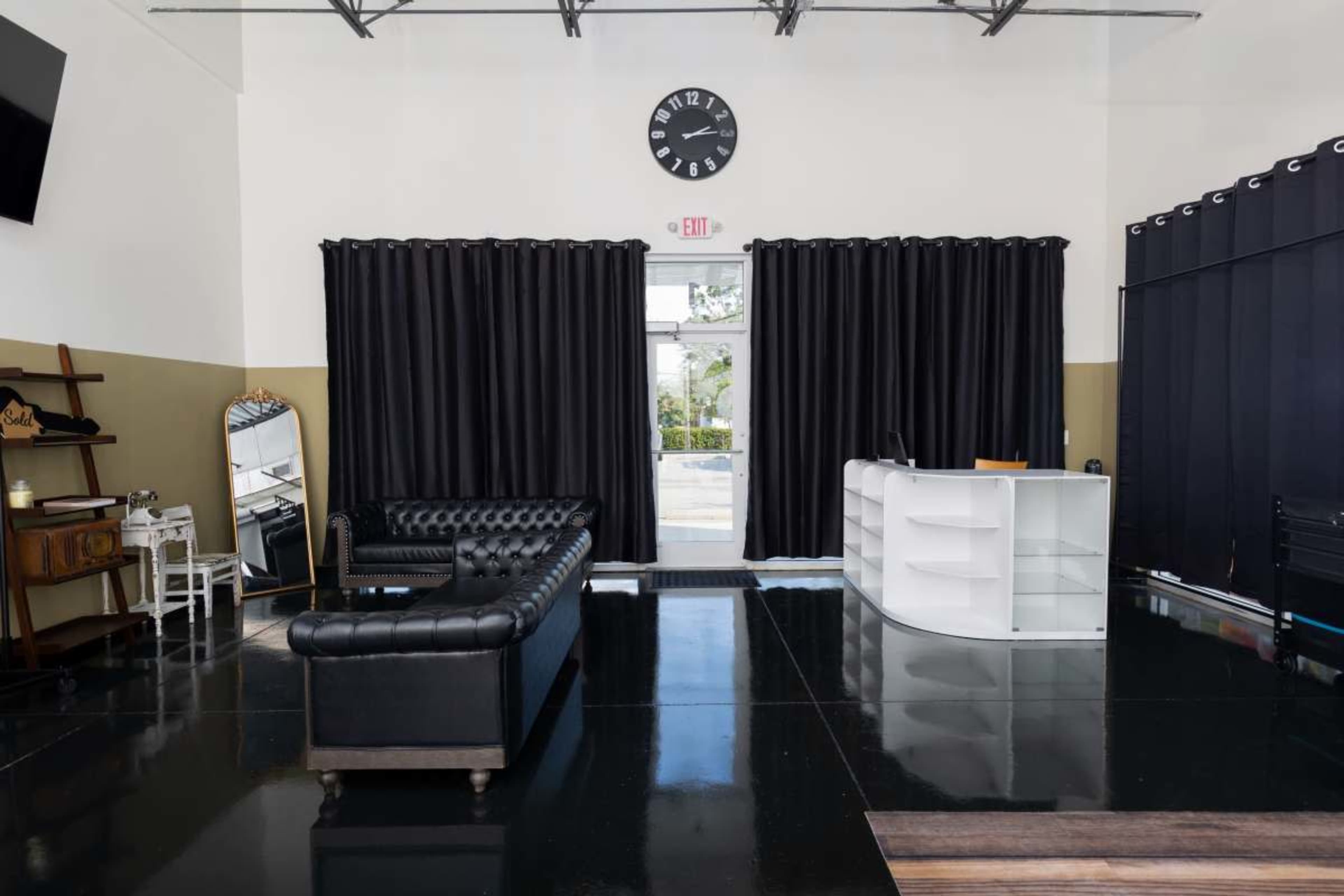 The image shows a spacious interior with a black leather couch, a white reception desk, large black curtains, and a clock on the wall.