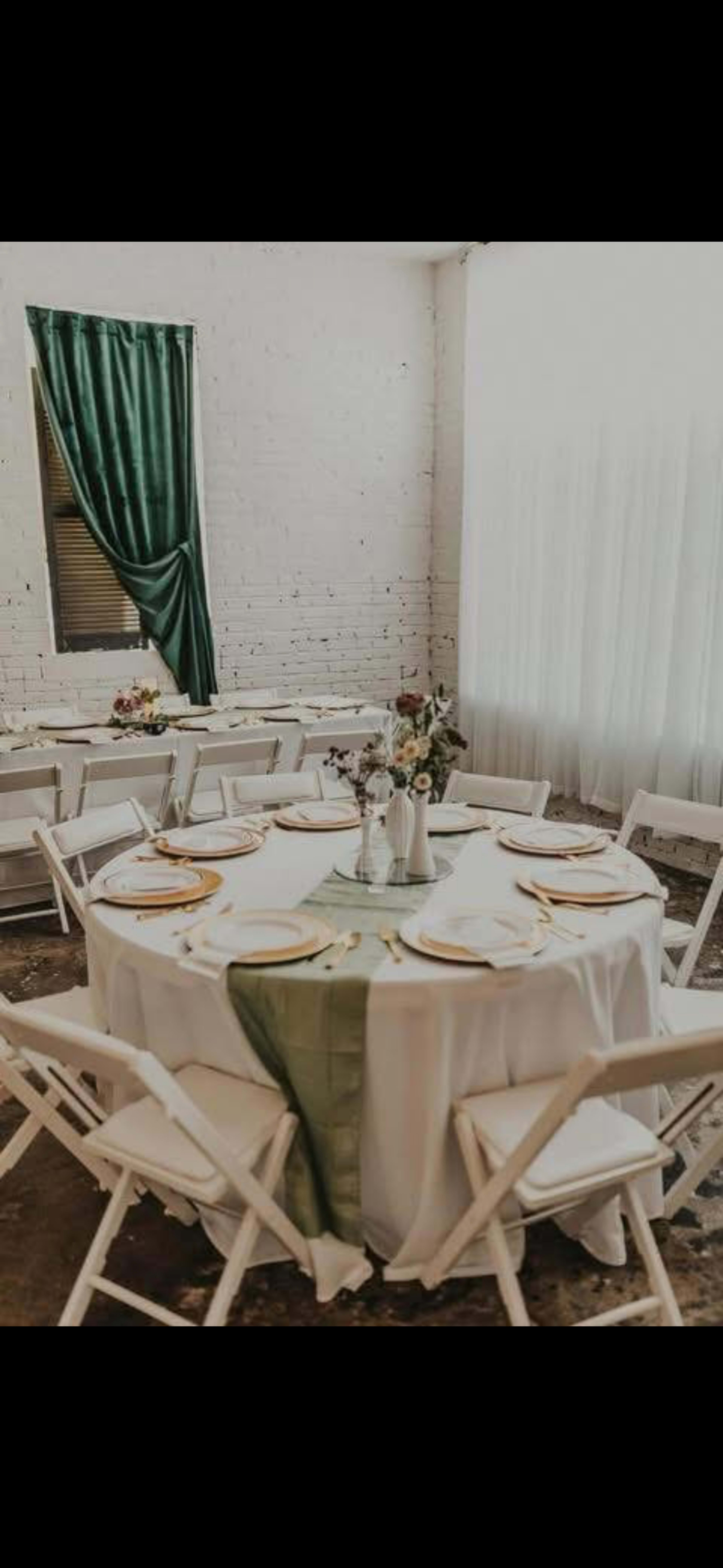A round table with white linens and green accents is elegantly set for a dining event, surrounded by white chairs in a well-lit room with a large window.