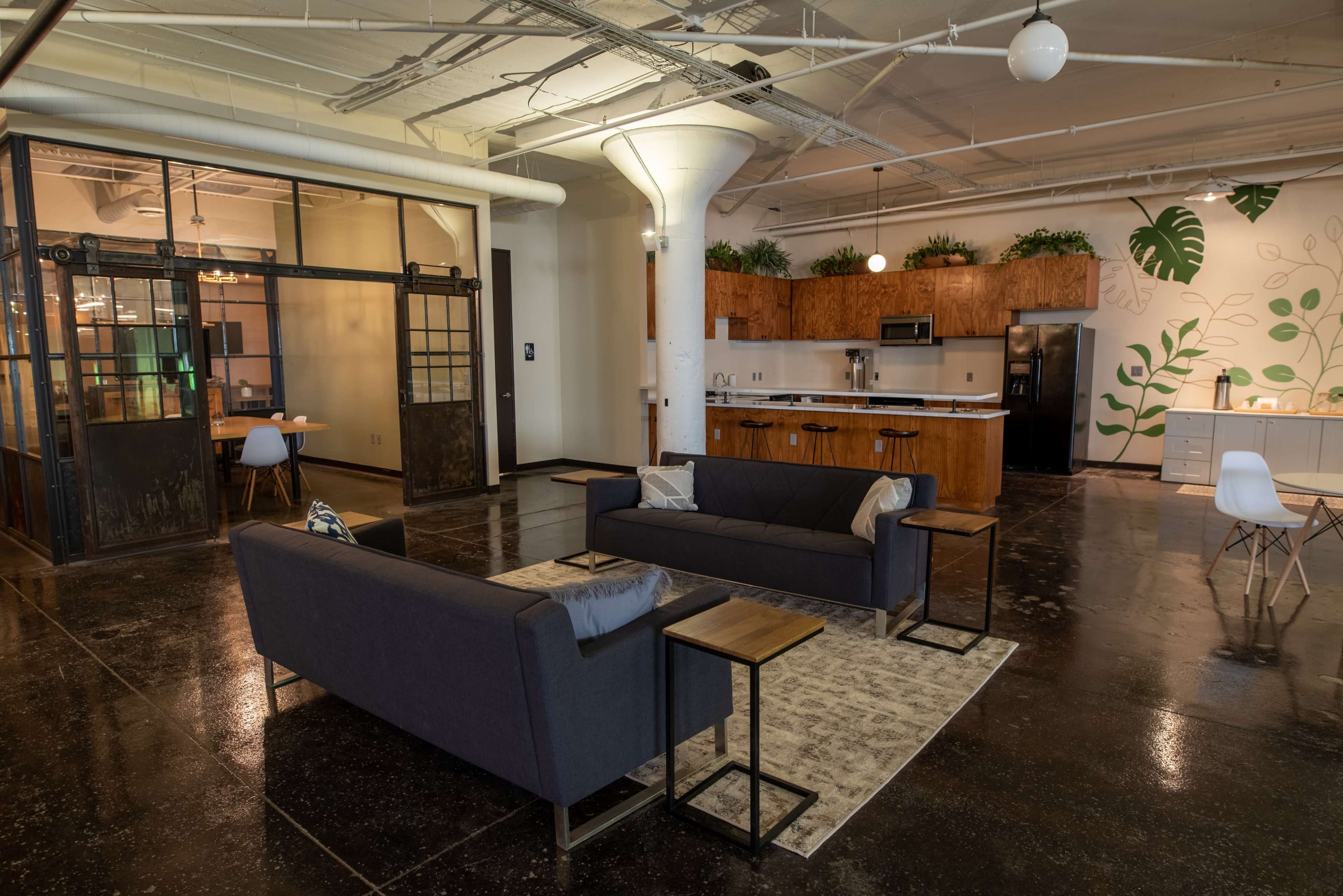 A modern, open-concept lounge area with two sofas, small side tables, and a kitchenette in the background, featuring wood cabinetry and plants.