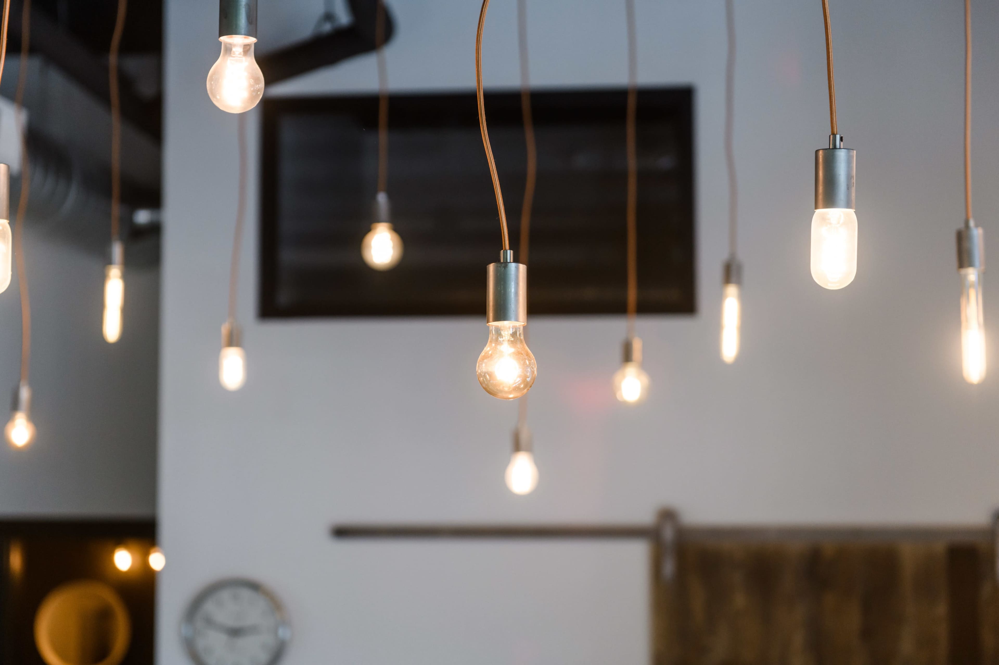 A group of hanging light bulbs with metal fixtures illuminates a room with a dark wall and a clock in the background.