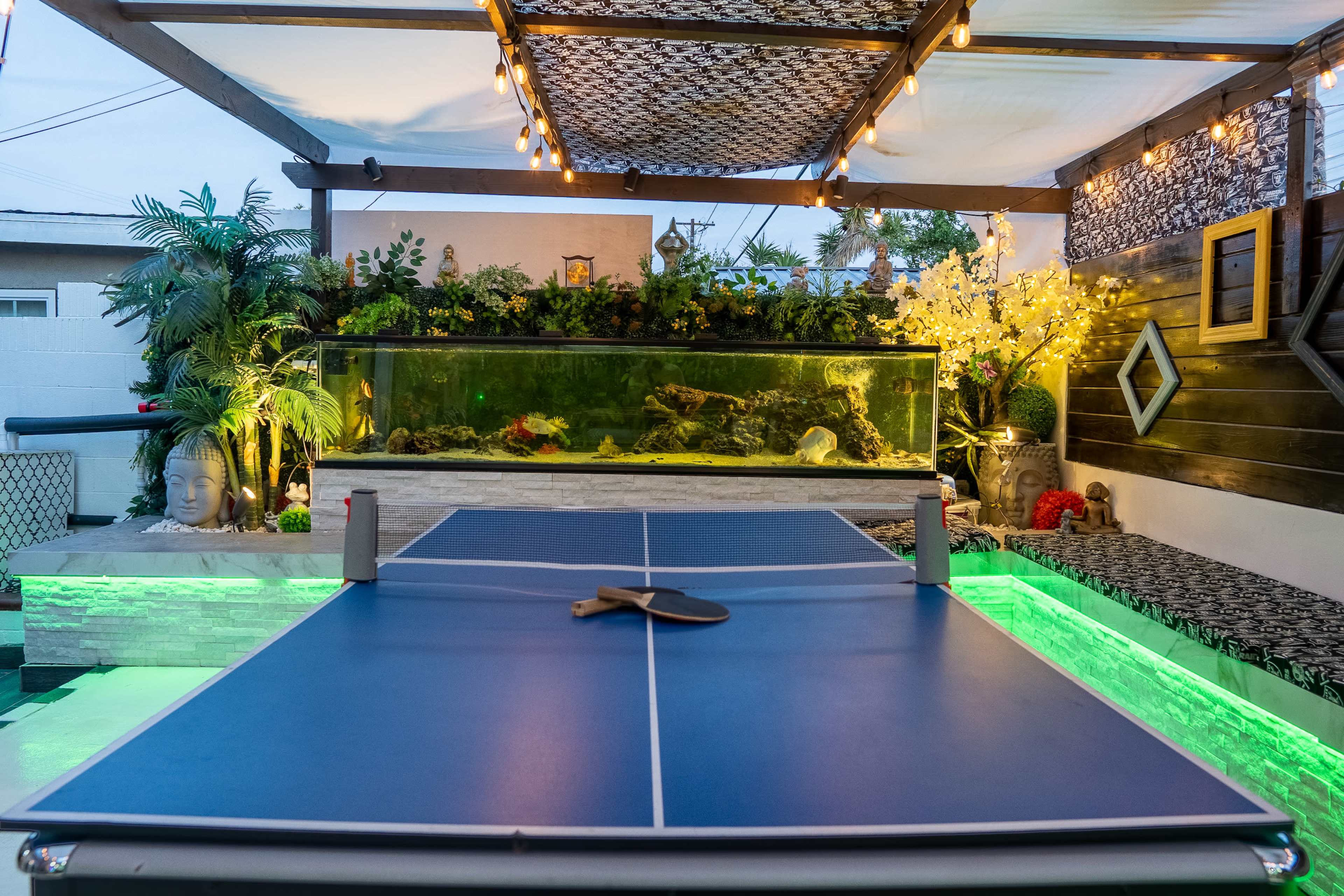 A backyard patio area with a blue ping pong table in the foreground, an aquarium filled with aquatic life in the background, and decorative lighting and plants around the space.