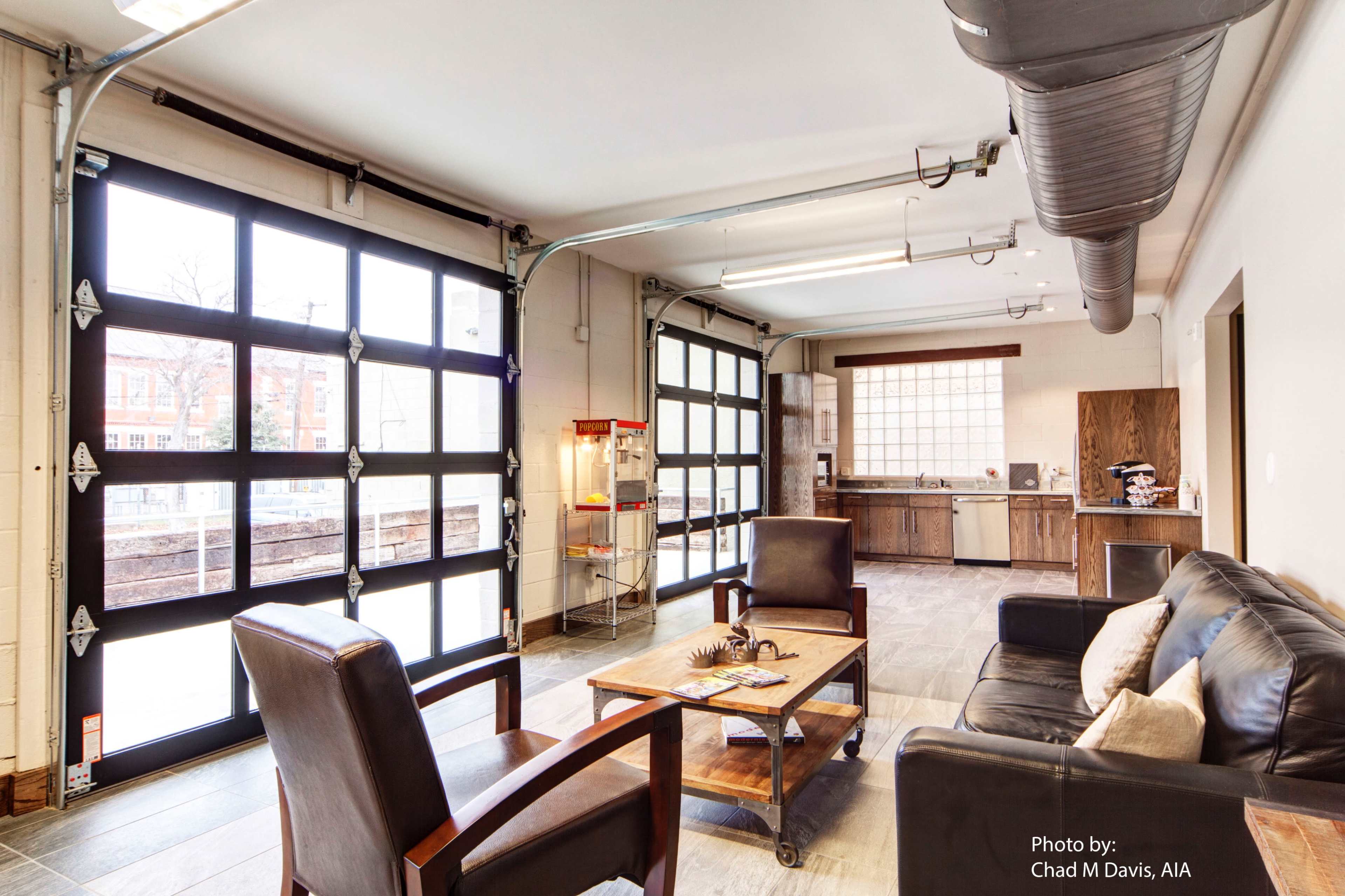 A spacious garage with large glass panel doors, a seating area featuring two armchairs, and a table in front of a kitchen space with wooden cabinetry.