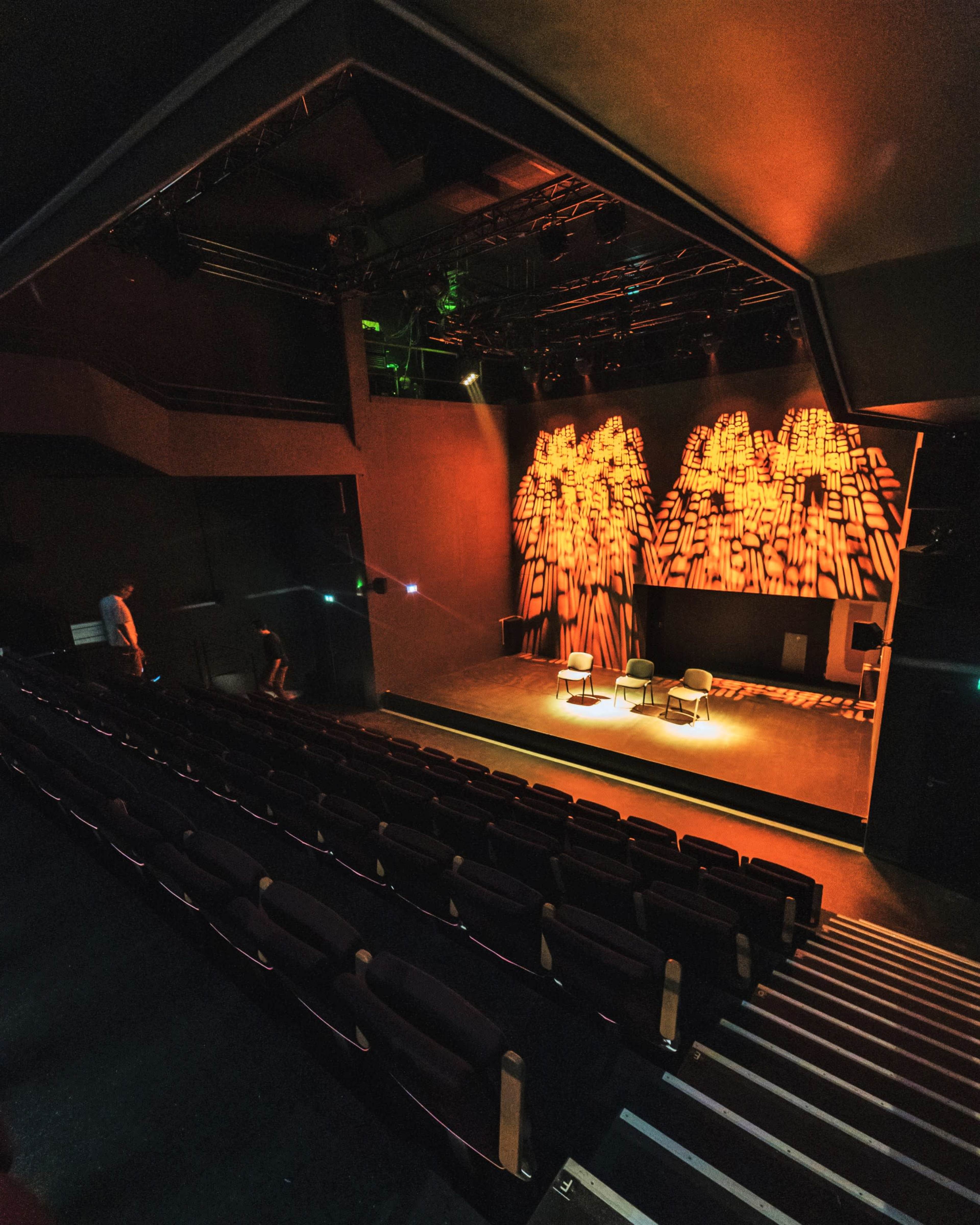 A theater stage with three chairs illuminated by warm lighting, surrounded by a dimly lit seating area.