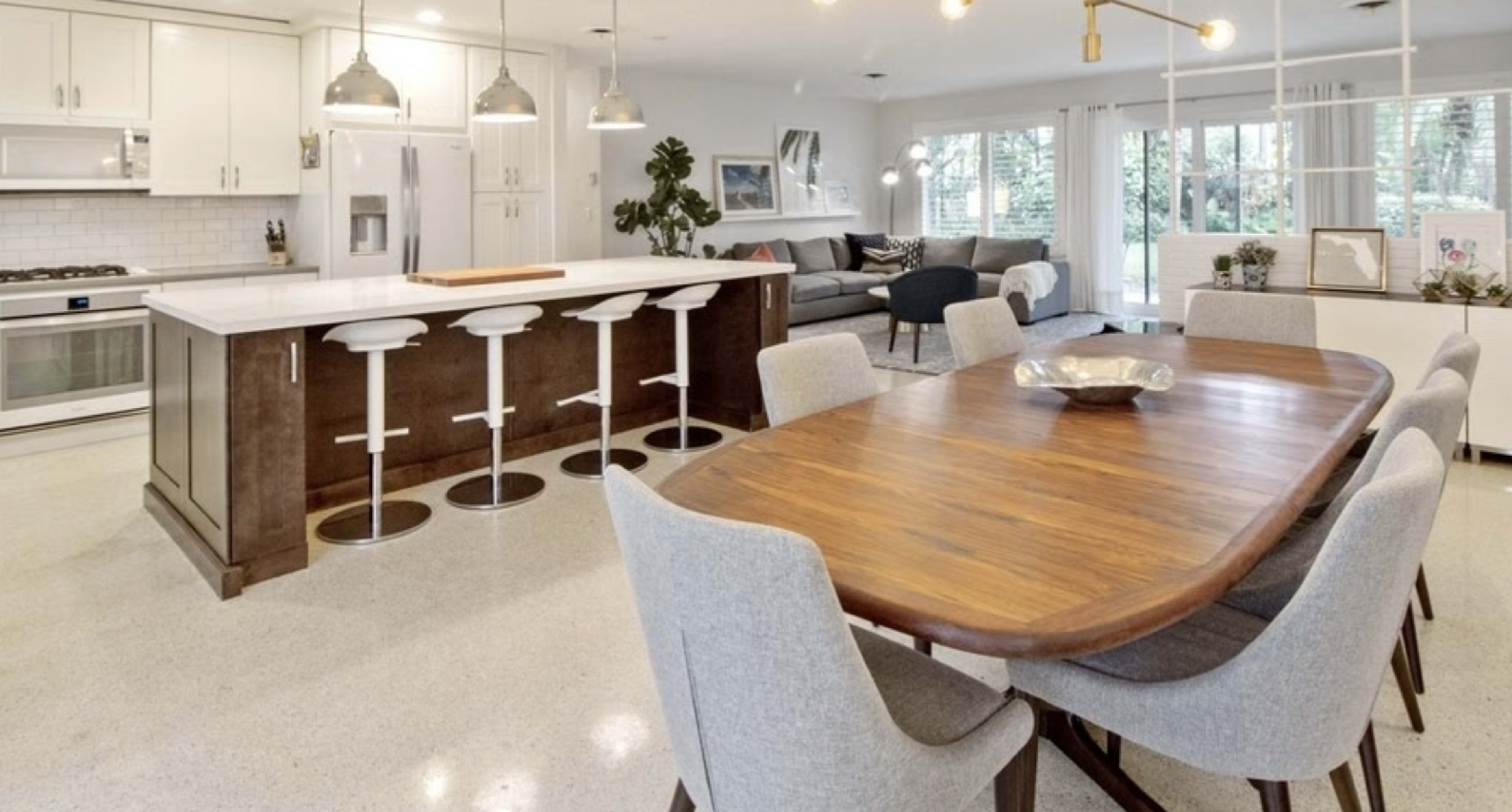The image shows a modern kitchen and dining area featuring a large wooden table surrounded by gray chairs, an island with bar stools, and a living room with a couch and large windows.