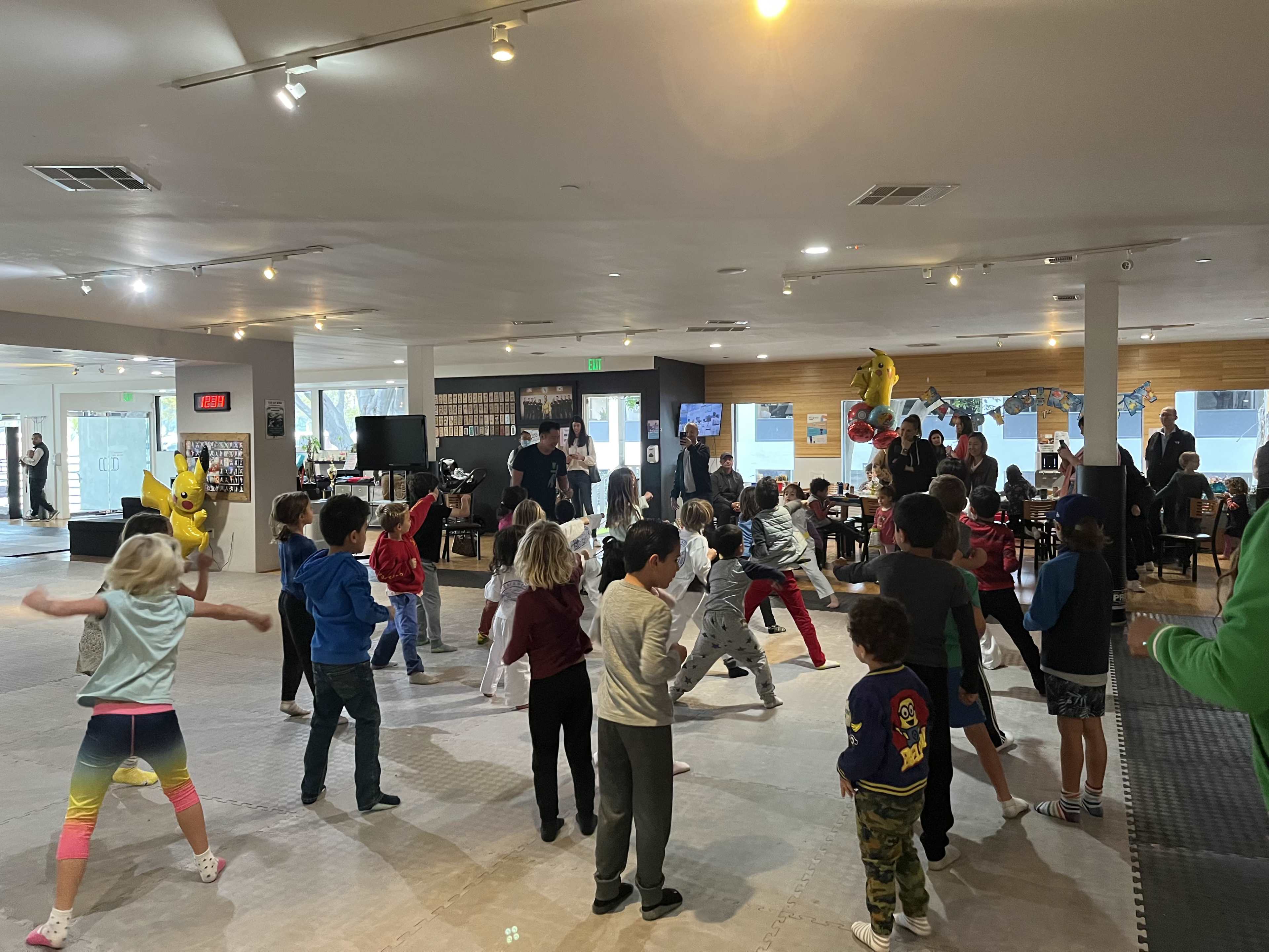 A group of children dance in a spacious indoor area filled with people and decorations.