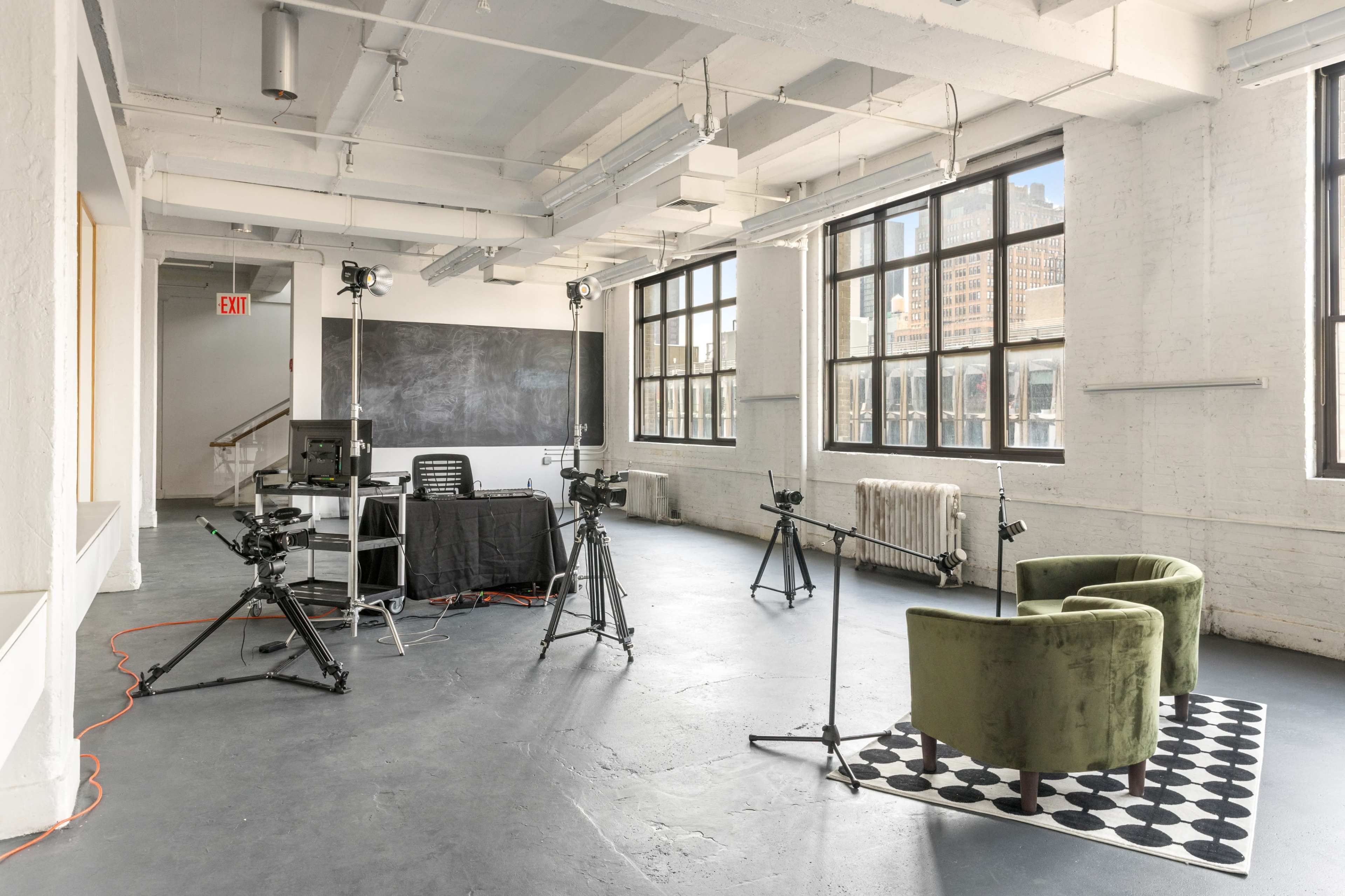 The image shows a spacious, well-lit studio with two green chairs, a black and white patterned rug, and several camera equipment setups facing the chairs.