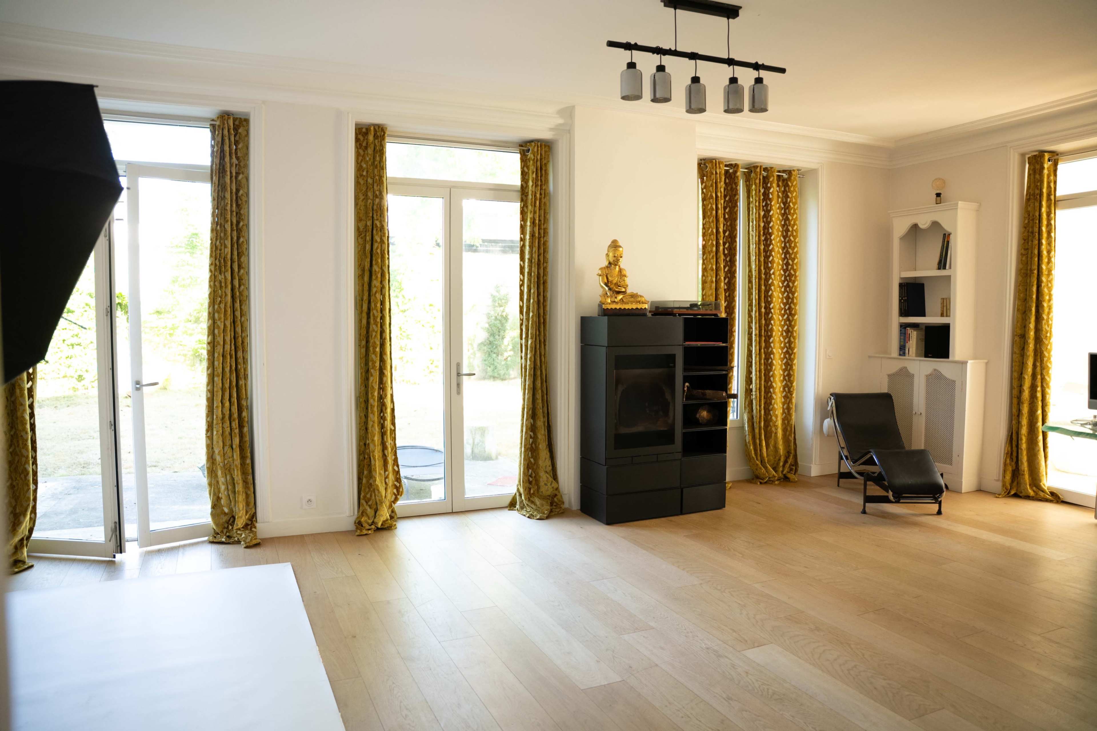 The image shows a spacious room with large windows, golden curtains, a black cabinet with a statue, and a lounge chair.