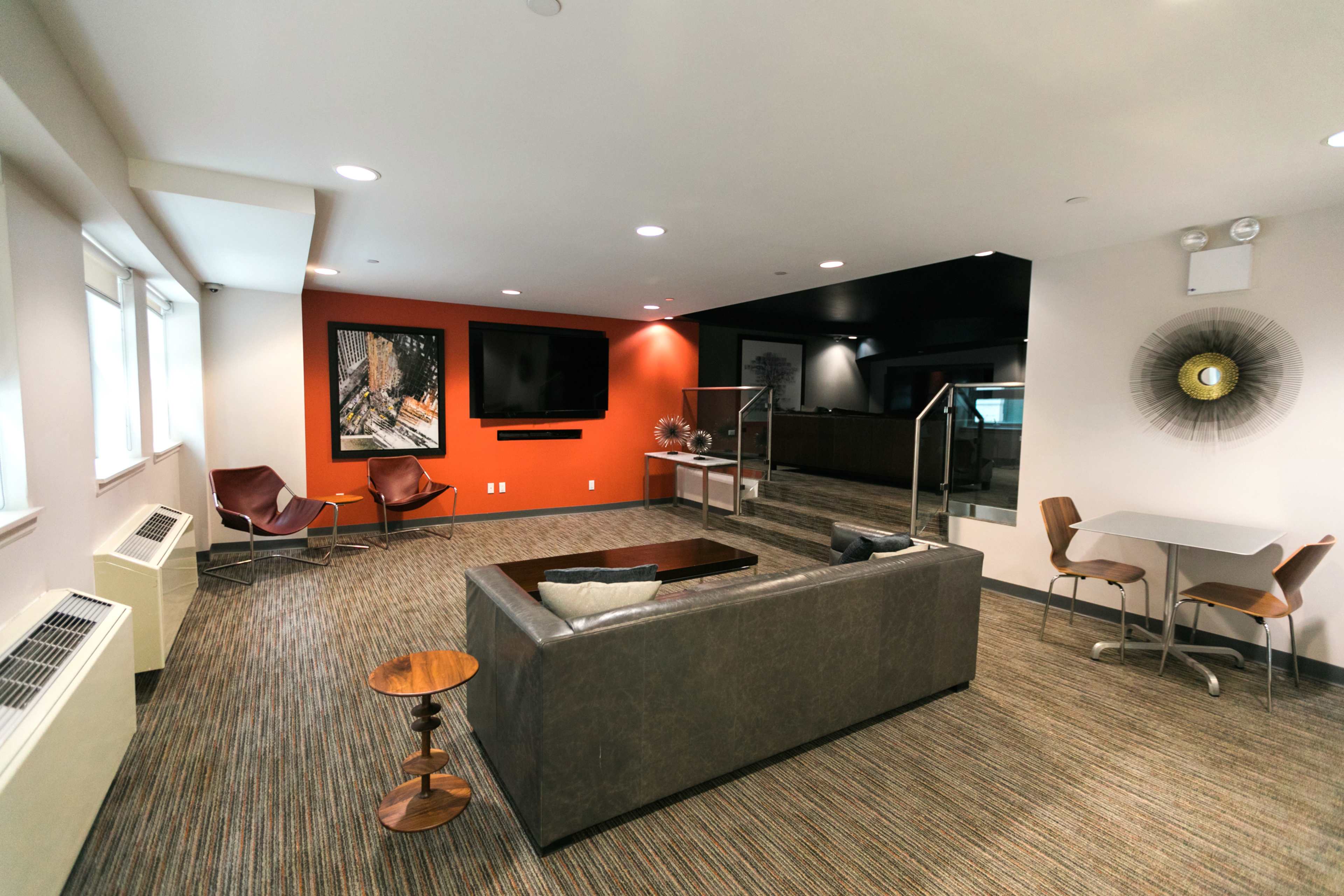 A modern lounge area with a gray sofa, two brown chairs, a side table, a flat-screen TV on the wall, and artwork, set in a well-lit space with varying wall colors.