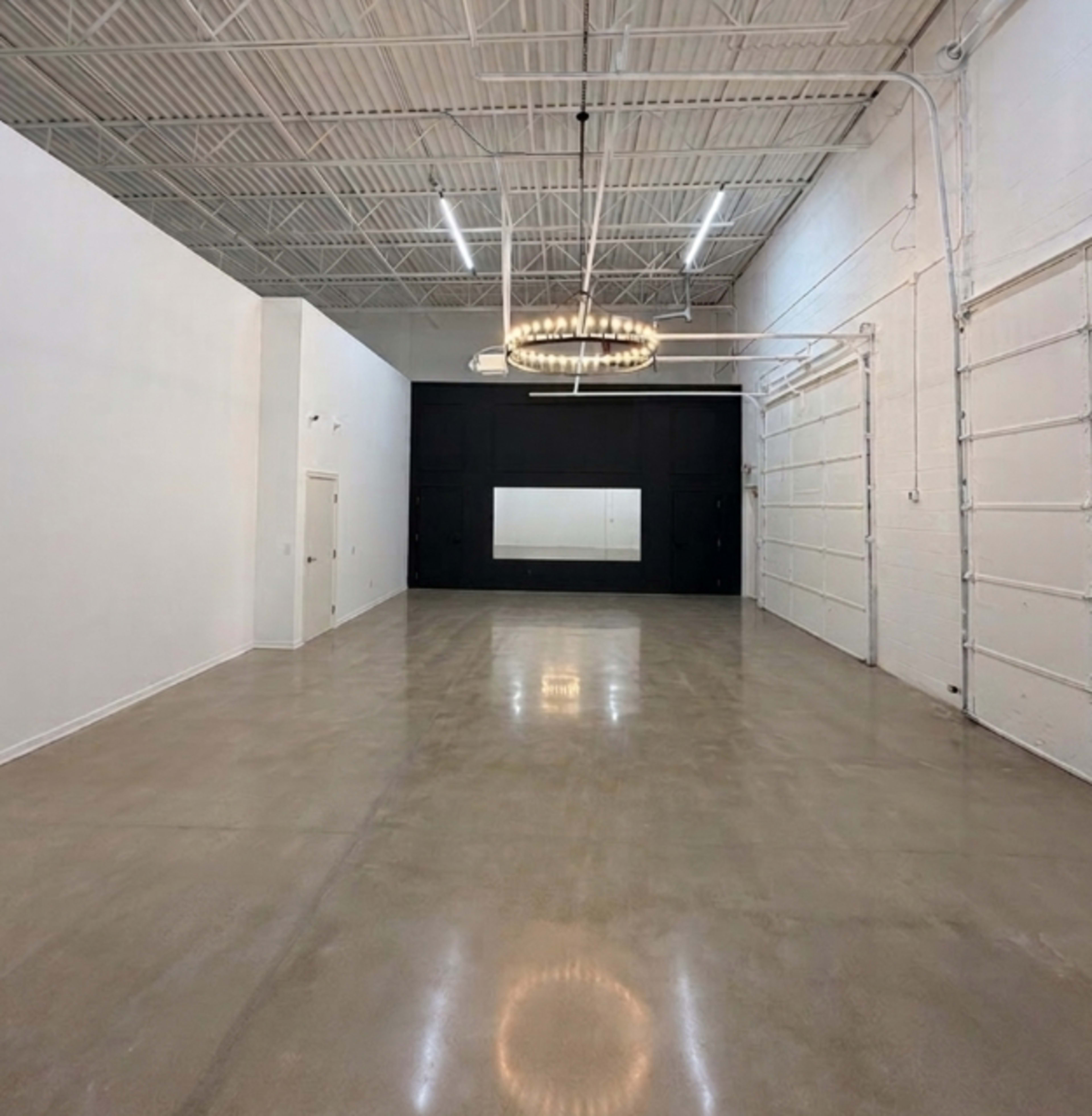 The image shows a spacious room with polished concrete flooring, white walls, a large black wall with a mirror, and a circular chandelier hanging from the ceiling.
