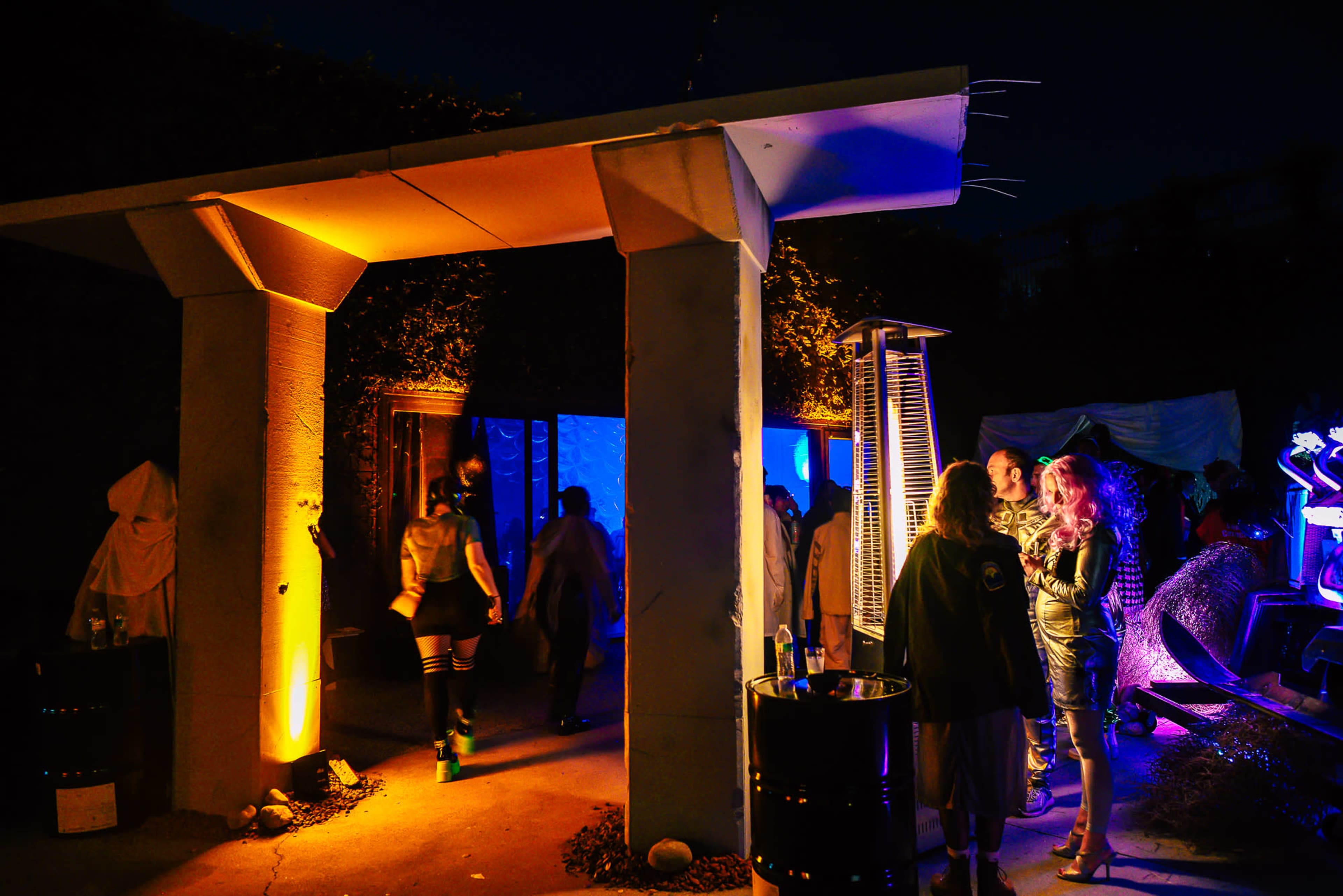 A crowd of people gathers outside a building at night, illuminated by colorful lights and heat lamps.