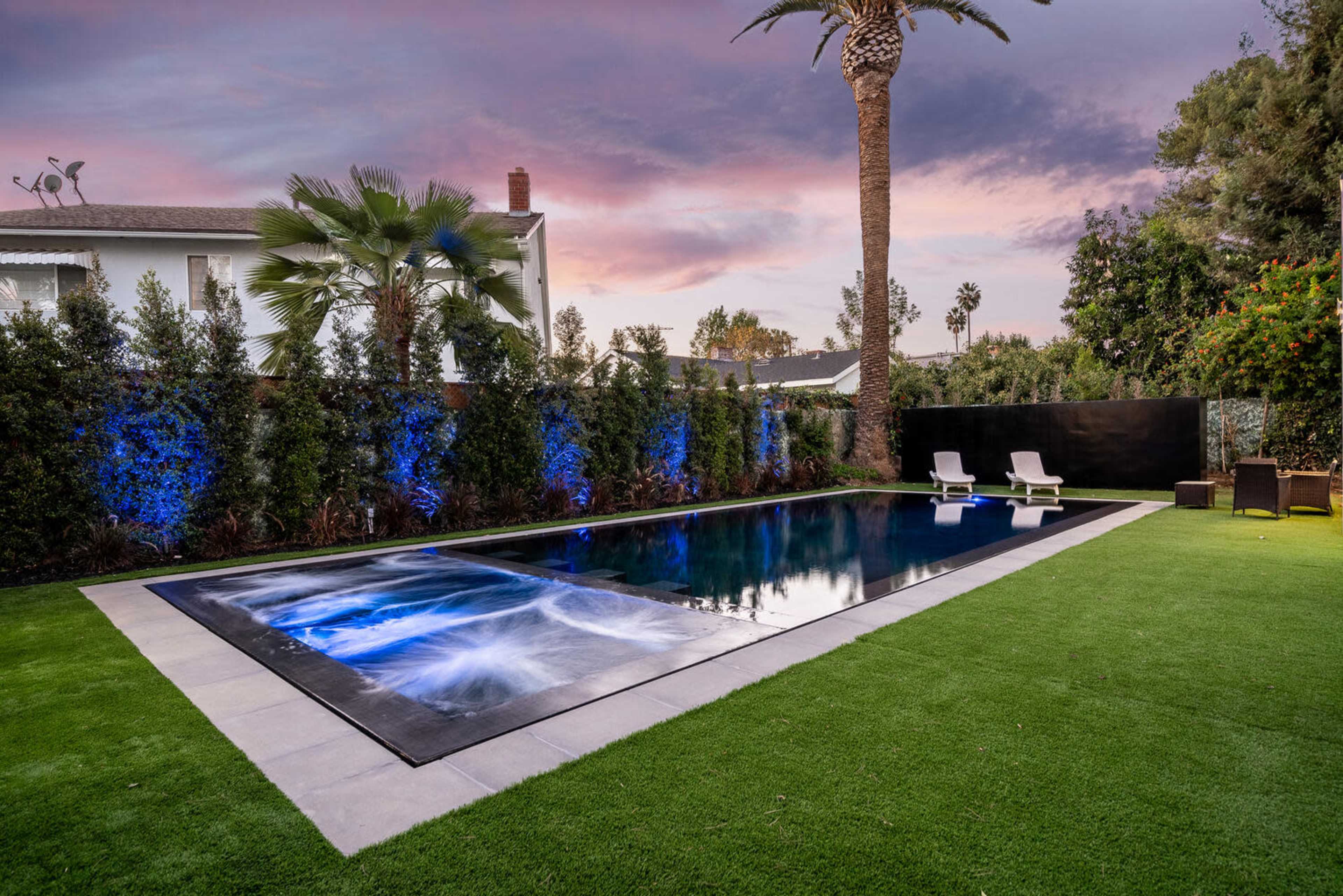 A rectangular swimming pool is framed by manicured grass and palm trees, with two white lounge chairs positioned nearby.