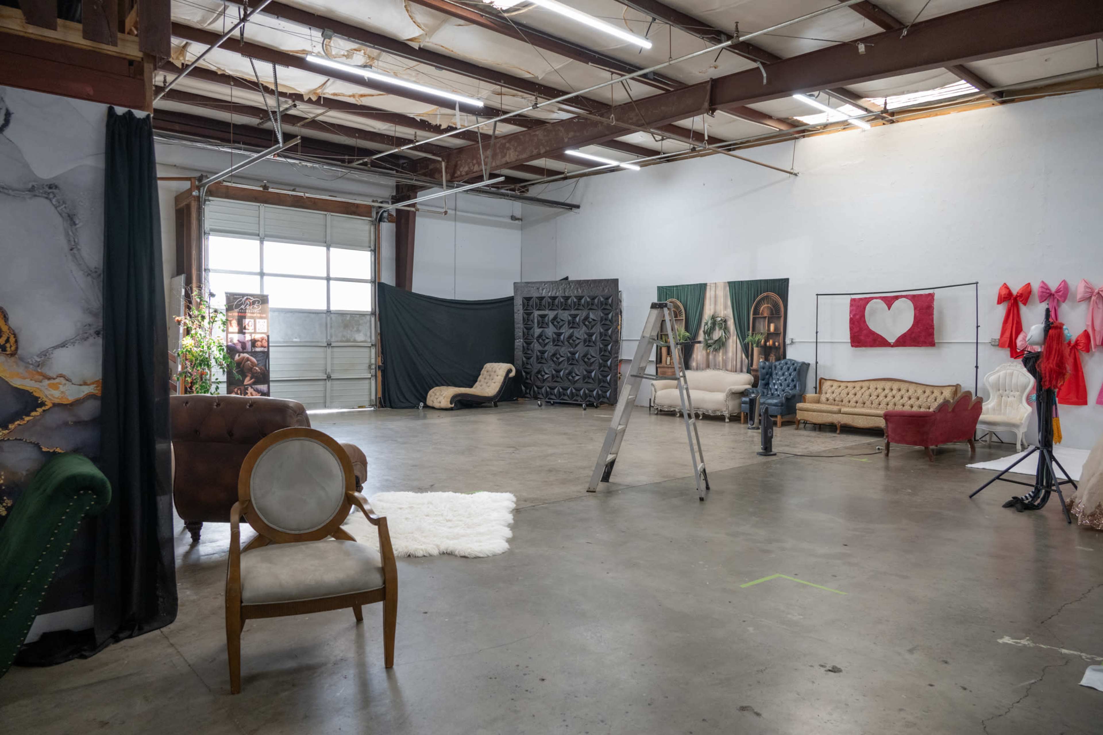 The image shows a spacious studio with various styled furniture pieces, a ladder, and several backdrop setups against the walls.