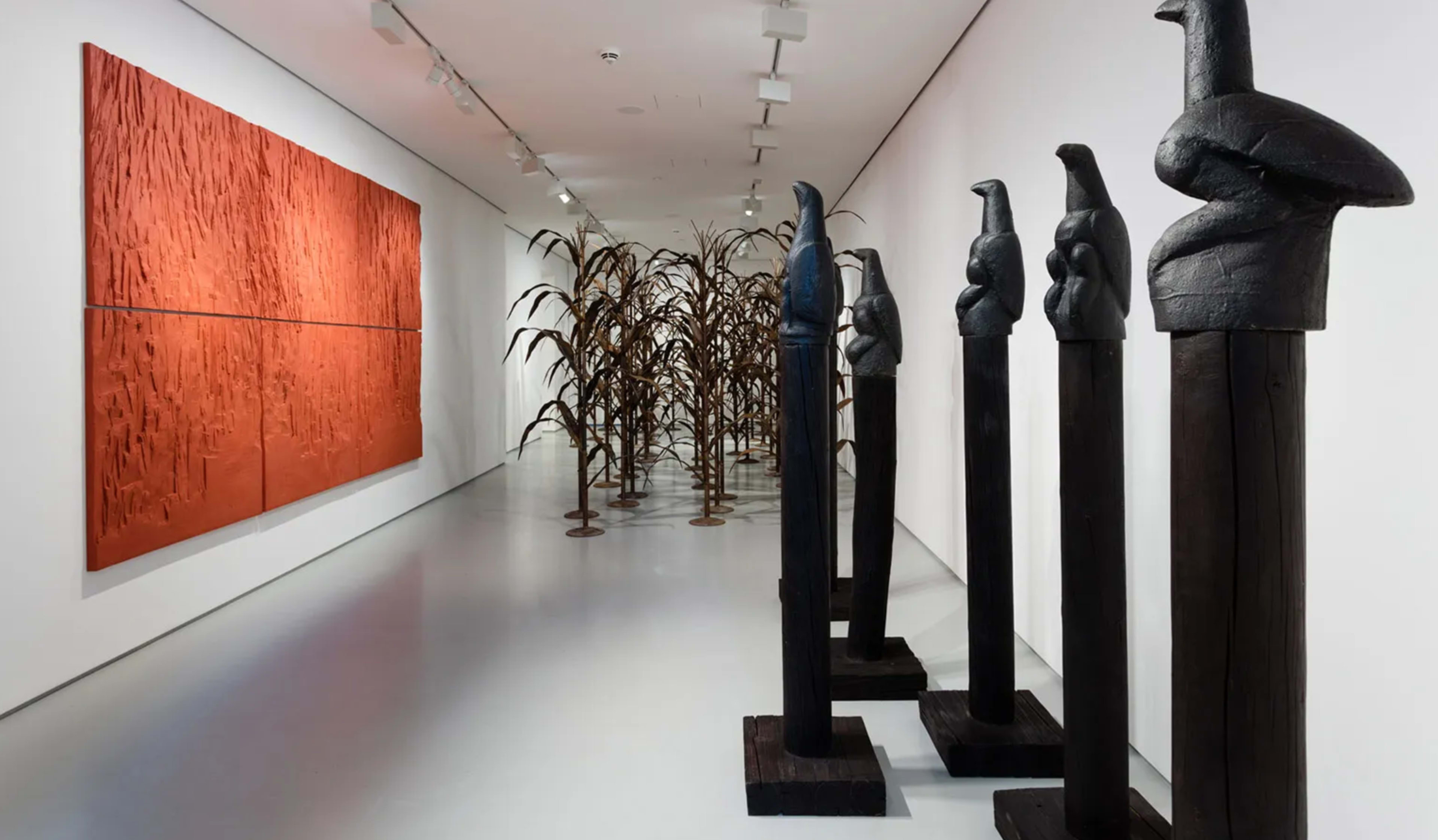 The image shows an art gallery featuring tall black sculptures arranged on the floor, a series of brown corn stalks in the middle, and a large textured orange artwork on the wall.