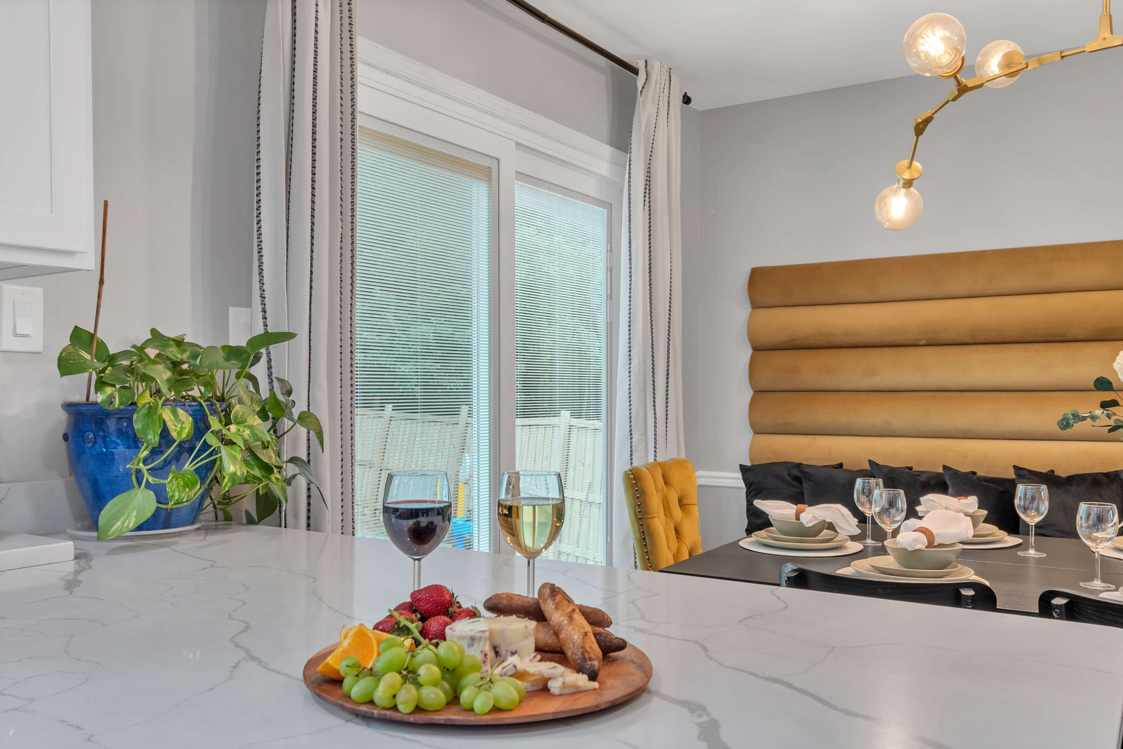 A wooden platter with assorted cheeses, fruits, and bread sits on a marble countertop in a modern kitchen, while a dining area with a yellow chair and large windows is visible in the background.