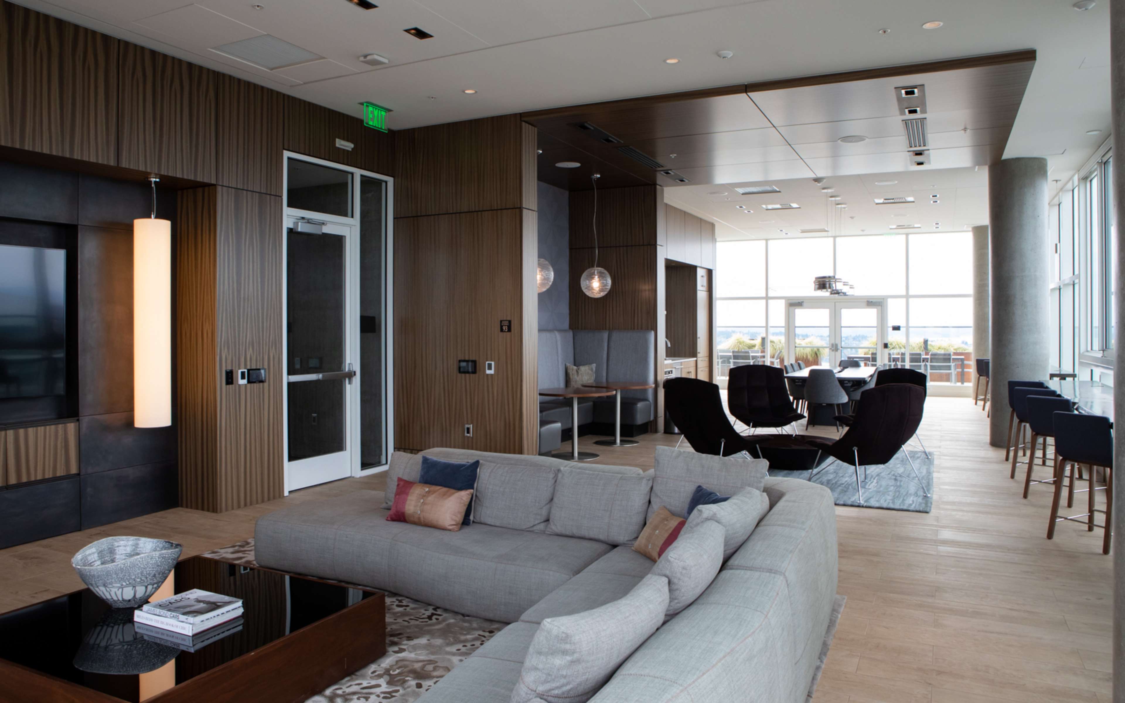 The image shows a modern living room with a spacious layout, featuring a sectional sofa, a coffee table, and a dining area with large windows that provide a view.