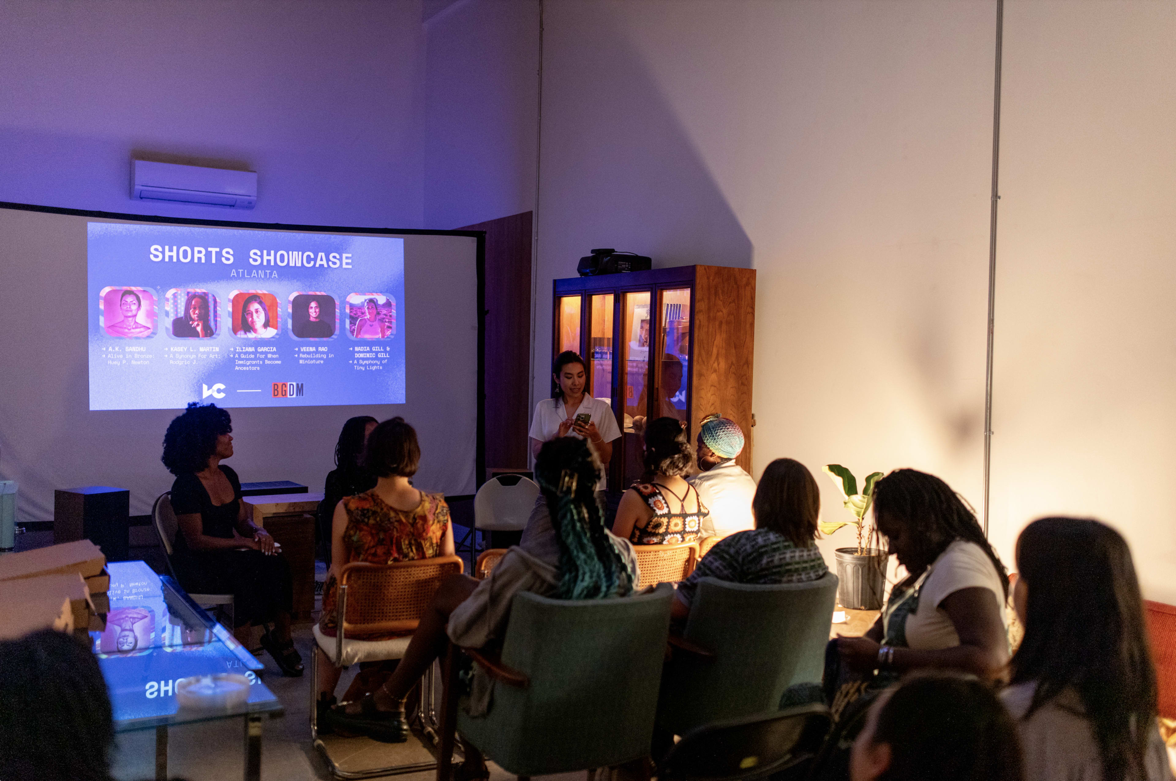 A group of people sits in a room facing a screen displaying information about a shorts showcase event in Atlanta, while a woman stands in front speaking to the audience.